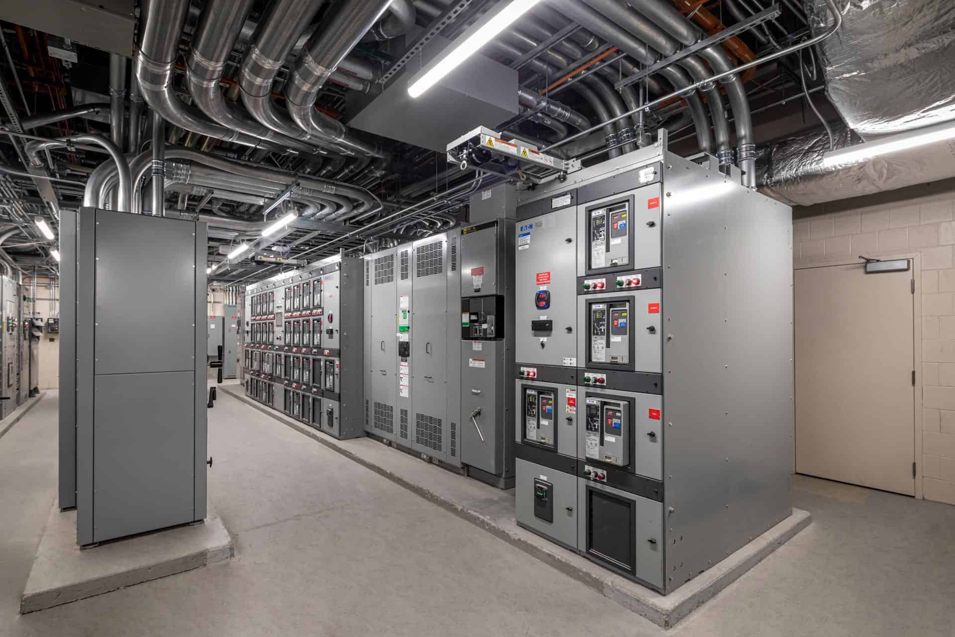 This image shows the electrical switchgear room at Riley Hospital for Children at IU Health Maternity Tower, featuring a row of large gray electrical distribution panels and control equipment. The room contains multiple electrical switchgear cabinets with various meters, switches, and control interfaces that manage the hospital's power distribution systems. An extensive network of conduits, pipes, and ductwork runs along the exposed ceiling, typical of a hospital's critical infrastructure areas. The space has concrete flooring with proper electrical grounding and safety measures, and includes emergency lighting systems. This electrical room is essential for providing reliable power distribution throughout the hospital facility, ensuring continuous operation of medical equipment and building systems vital to patient care.