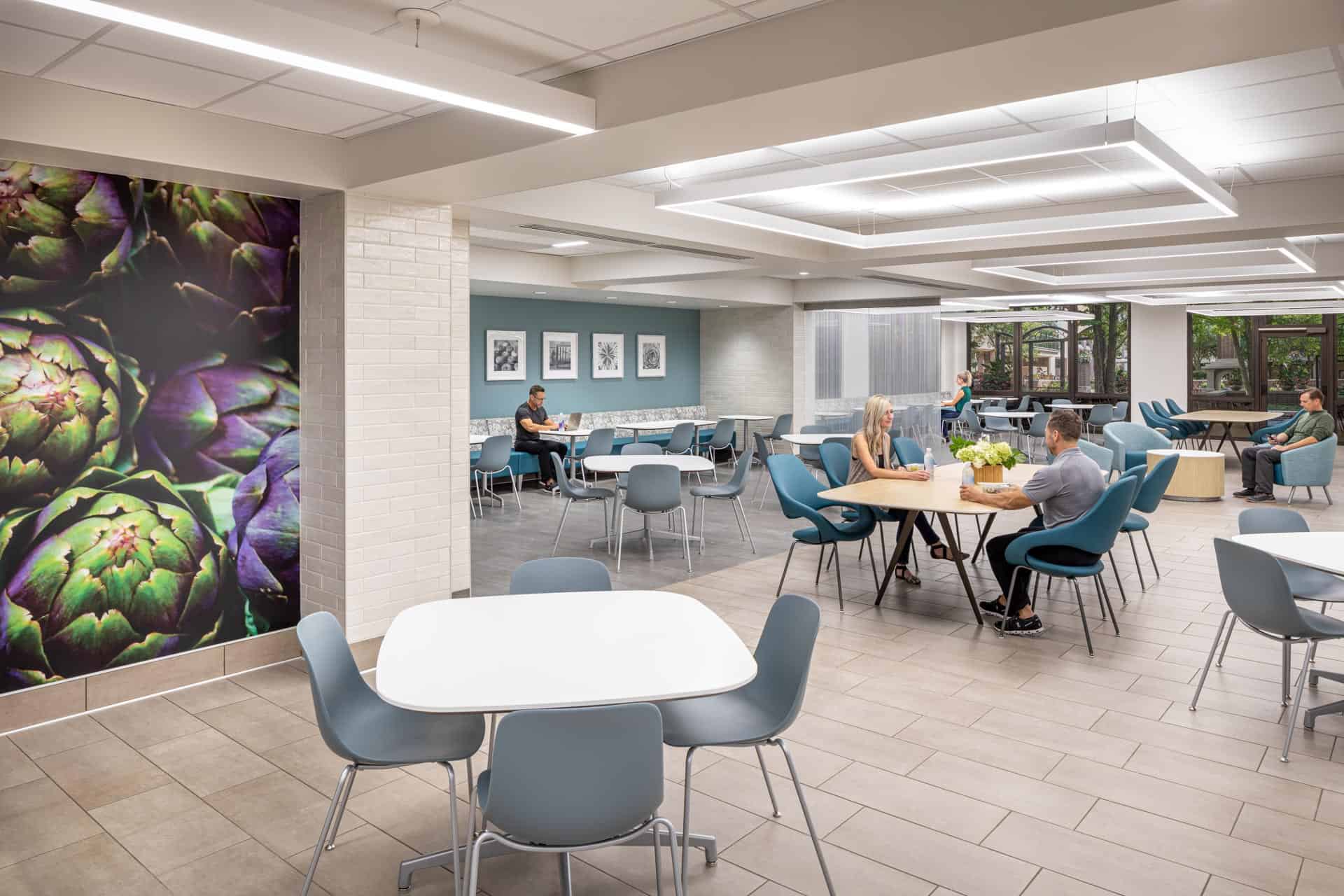 This image shows a modern cafeteria or dining area at Riley Hospital for Children at IU Health Maternity Tower with a contemporary design featuring various seating arrangements. The space includes round and rectangular tables with teal and gray chairs, accommodating both small groups and individuals. A striking large-scale botanical wall graphic depicting purple artichokes serves as an artistic focal point on the left wall. The dining area has an open layout with geometric ceiling details, large windows providing natural light and views of greenery outside, and a teal accent wall with framed artwork. The space is designed to create a comfortable, restaurant-like atmosphere for patients, families, and hospital staff to dine and socialize.