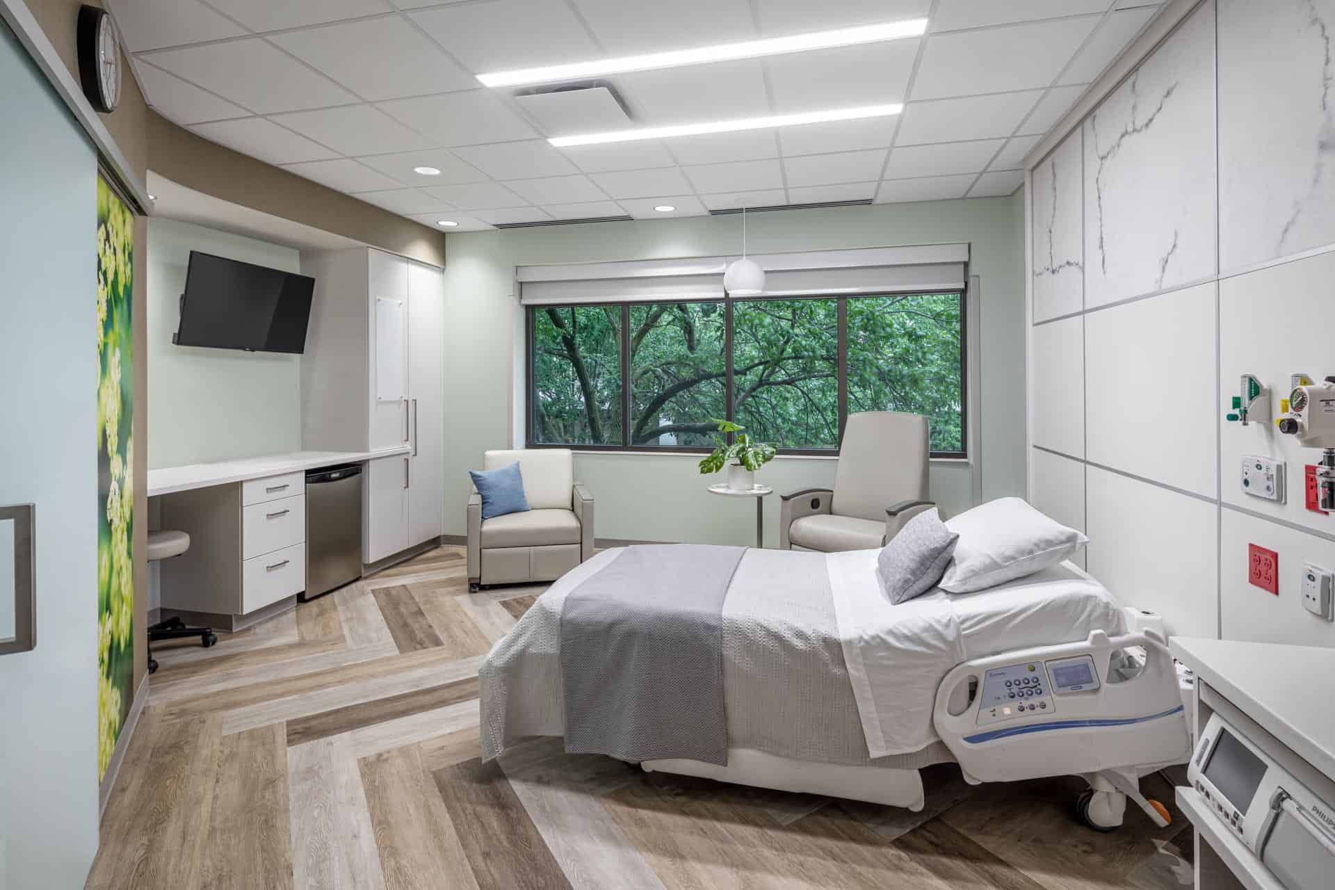 This image shows a modern, well-appointed maternity room at Riley Hospital for Children at IU Health Maternity Tower. The spacious room features a hospital bed with white linens and gray accents, positioned near a large window overlooking lush green trees. The room includes comfortable seating with two beige armchairs and a small side table, along with built-in white cabinetry, a wall-mounted television, and wood-look flooring. The clean, contemporary design incorporates calming neutral tones with white walls, marble-pattern wall panels, and a colorful abstract artwork, creating a home-like atmosphere for new families while maintaining the functionality of a medical facility.