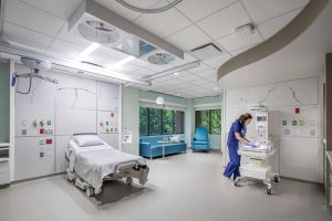 This image shows a modern labor and delivery room at Riley Hospital for Children at IU Health Maternity Tower designed for medical procedures and patient comfort. The room features a hospital bed with overhead surgical lighting and medical gas outlets mounted on the wall, while a healthcare provider in blue scrubs tends to a newborn in a mobile bassinet. The space includes a comfortable blue sofa and chair positioned near large windows that provide natural light and views of greenery outside. The room has a clean, clinical design with white and soft green color schemes, specialized medical equipment integrated into the ceiling and walls, and marble-patterned accent panels that create a calming yet fully functional environment for childbirth and immediate newborn care.