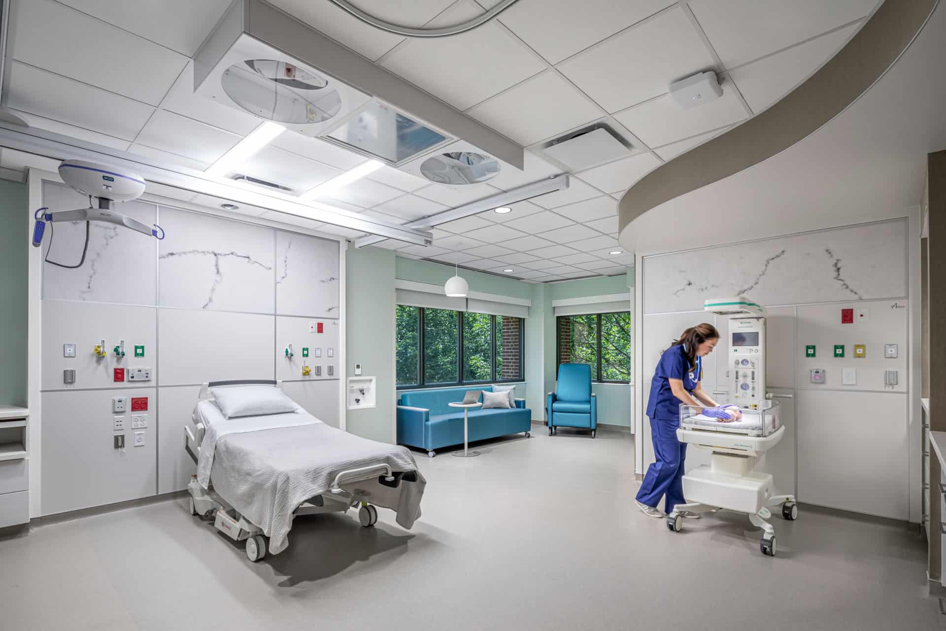 This image shows a modern labor and delivery room at Riley Hospital for Children at IU Health Maternity Tower designed for medical procedures and patient comfort. The room features a hospital bed with overhead surgical lighting and medical gas outlets mounted on the wall, while a healthcare provider in blue scrubs tends to a newborn in a mobile bassinet. The space includes a comfortable blue sofa and chair positioned near large windows that provide natural light and views of greenery outside. The room has a clean, clinical design with white and soft green color schemes, specialized medical equipment integrated into the ceiling and walls, and marble-patterned accent panels that create a calming yet fully functional environment for childbirth and immediate newborn care.