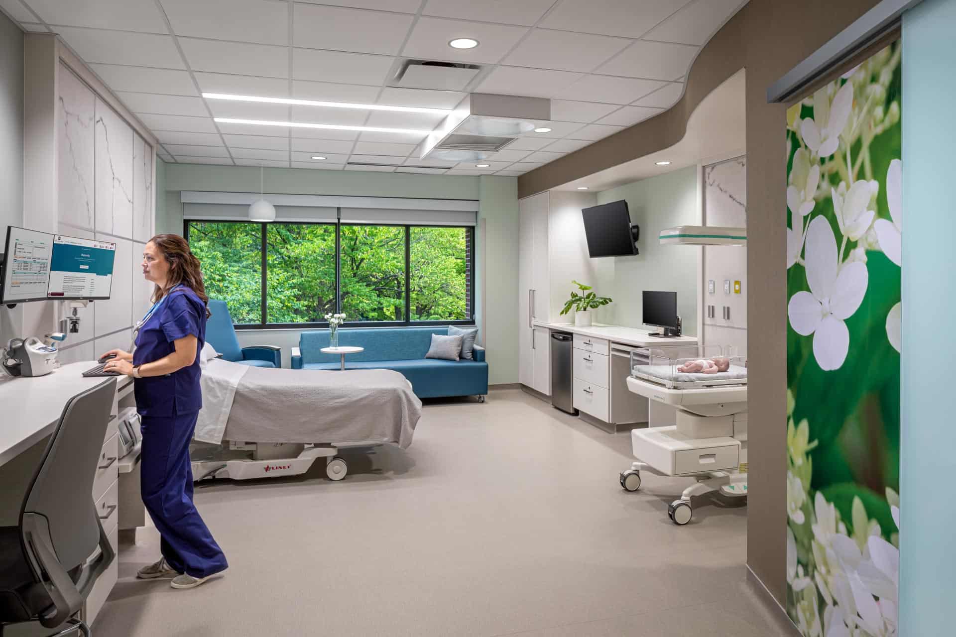 This image shows a modern labor and delivery room at Riley Hospital for Children at IU Health Maternity Tower where a healthcare provider in blue scrubs is working at a computer station. The room features a hospital bed positioned near large windows with views of greenery, creating a calming natural environment. A blue sofa provides comfortable seating for family members, while medical equipment including monitors and a newborn care station are seamlessly integrated into the space. The room displays large-scale floral artwork featuring white blossoms on a green background, contributing to a soothing, home-like atmosphere. White cabinetry and modern finishes throughout the room combine medical functionality with patient comfort during the birthing process.