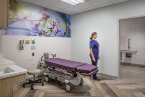 A healthcare professional in blue scrubs moves through a modern patient room at Riley Hospital for Children's Maternity Tower, featuring a purple-cushioned medical bed positioned beneath a large nature-themed wall mural of flowering branches. The room includes wood-look flooring, medical outlets and equipment mounted on the wall, a sink area with wood cabinetry, and an accessible bathroom, all designed to create a calming healing environment for maternity patients and their families.