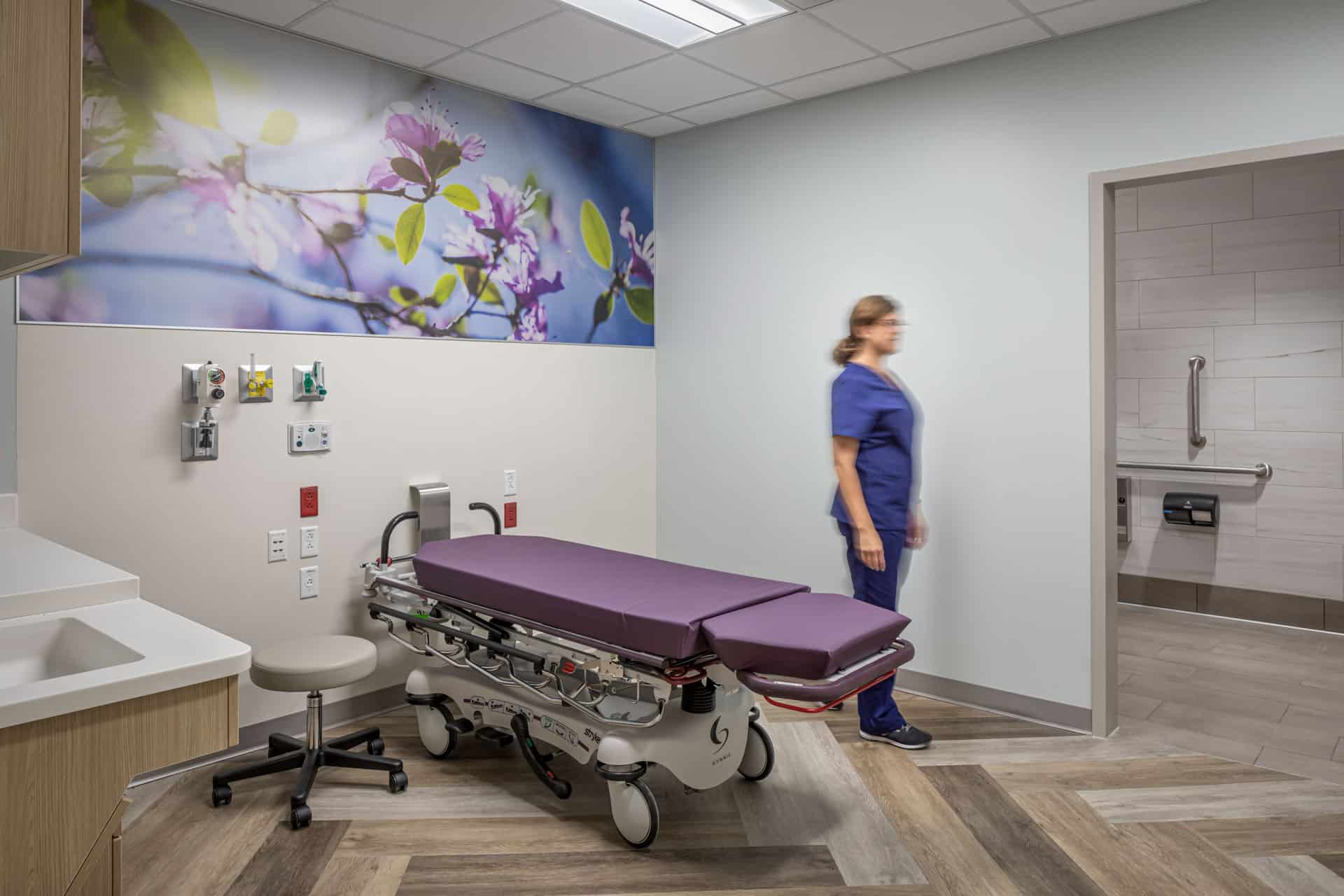 A healthcare professional in blue scrubs moves through a modern patient room at Riley Hospital for Children's Maternity Tower, featuring a purple-cushioned medical bed positioned beneath a large nature-themed wall mural of flowering branches. The room includes wood-look flooring, medical outlets and equipment mounted on the wall, a sink area with wood cabinetry, and an accessible bathroom, all designed to create a calming healing environment for maternity patients and their families.