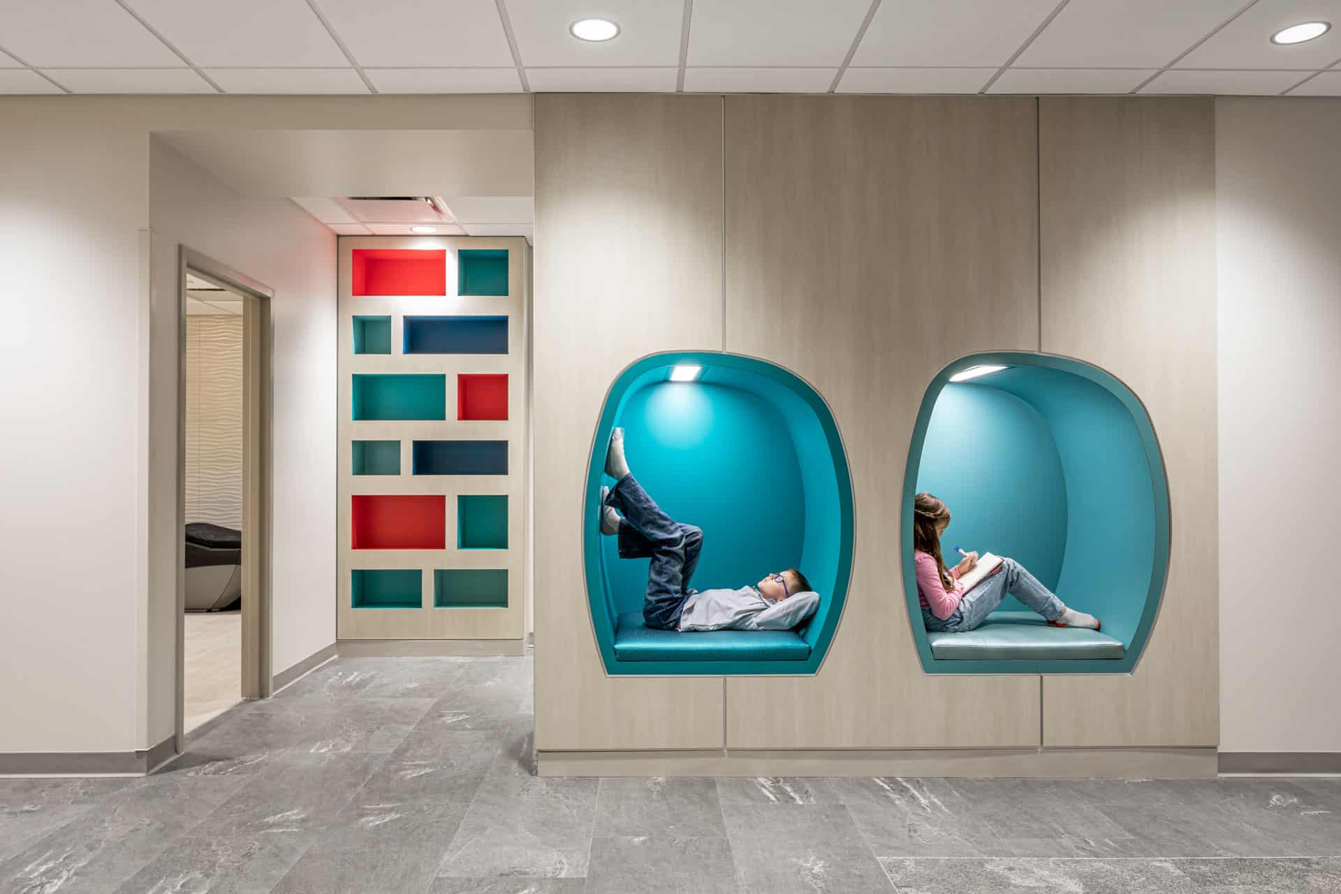 This image shows a modern hospital interior at Riley Hospital for Children at IU Health Maternity Tower featuring two distinctive turquoise-colored pod-style seating alcoves built into the wall. One pod contains a child lying down, while the other has a child sitting and reading. The space has a contemporary design with neutral beige and gray tones, polished concrete floors, and a colorful geometric art installation on the left wall featuring red, teal, and navy rectangular panels. The area includes recessed lighting in the drop ceiling and appears to be a comfortable waiting or quiet space designed for families and patients.