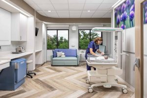 This image shows a modern maternity room at Riley Hospital for Children at IU Health Maternity Tower where a healthcare provider in blue scrubs is caring for a newborn in a hospital bassinet. The room features a contemporary design with white cabinetry, herringbone wood-look flooring, and large windows overlooking greenery. A light blue sofa with decorative pillows provides seating for family members, while purple floral artwork on the wall adds color to the space. The room includes medical equipment, built-in storage, and is designed to create a comfortable, home-like environment for new mothers and their babies during their hospital stay.