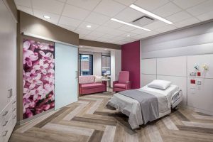 This image shows a modern patient room at Riley Hospital for Children at IU Health Maternity Tower featuring a hospital bed with gray bedding positioned near a window. The room has a sophisticated design with herringbone wood-look flooring and a striking large-scale floral artwork depicting pink blossoms that serves as a focal point on one wall. A burgundy accent wall complements pink seating furniture including a sofa and chair, creating a warm and welcoming atmosphere. The space includes built-in white cabinetry for storage, medical outlets on the wall, and is designed to provide comfort for patients and their families while maintaining necessary medical functionality.