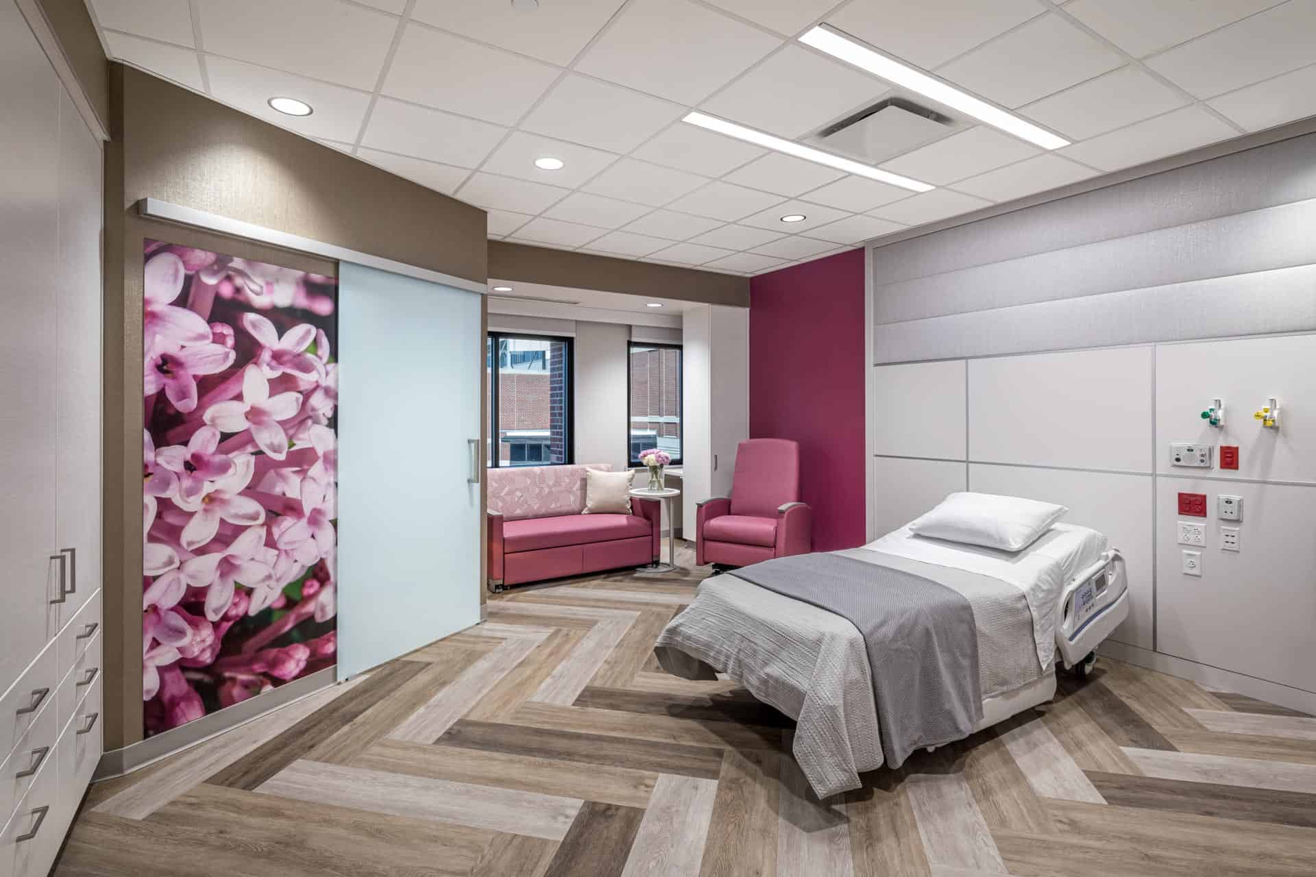 This image shows a modern patient room at Riley Hospital for Children at IU Health Maternity Tower featuring a hospital bed with gray bedding positioned near a window. The room has a sophisticated design with herringbone wood-look flooring and a striking large-scale floral artwork depicting pink blossoms that serves as a focal point on one wall. A burgundy accent wall complements pink seating furniture including a sofa and chair, creating a warm and welcoming atmosphere. The space includes built-in white cabinetry for storage, medical outlets on the wall, and is designed to provide comfort for patients and their families while maintaining necessary medical functionality.