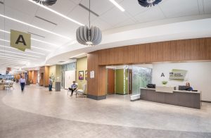 Converted mall interior now serving as a healthcare complex. A spacious lobby with modern design features linear lighting, a large decorative pendant light, and wood-paneled walls. Wayfinding signs marked 'A' and 'B' guide visitors. A reception desk is staffed, and several people sit or walk through the open, bright space with neutral-toned flooring and seating areas.