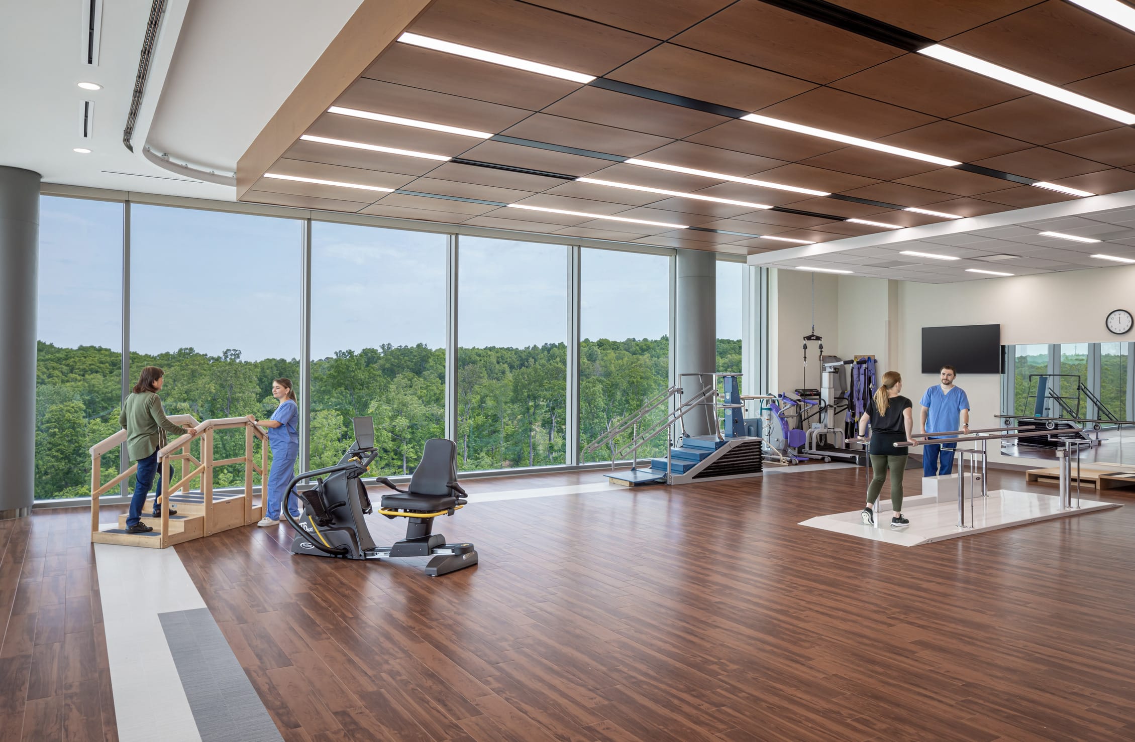 UNC Hospitals Hillsborough spacious rehabilitation room, featuring physical therapy equipment. The room is mostly empty and has sleek wooden floors. All of the walls are made of windows, offering a view of the lush trees outside. This image showcases BSA's expertise in designing spaces that prioritize patient care.