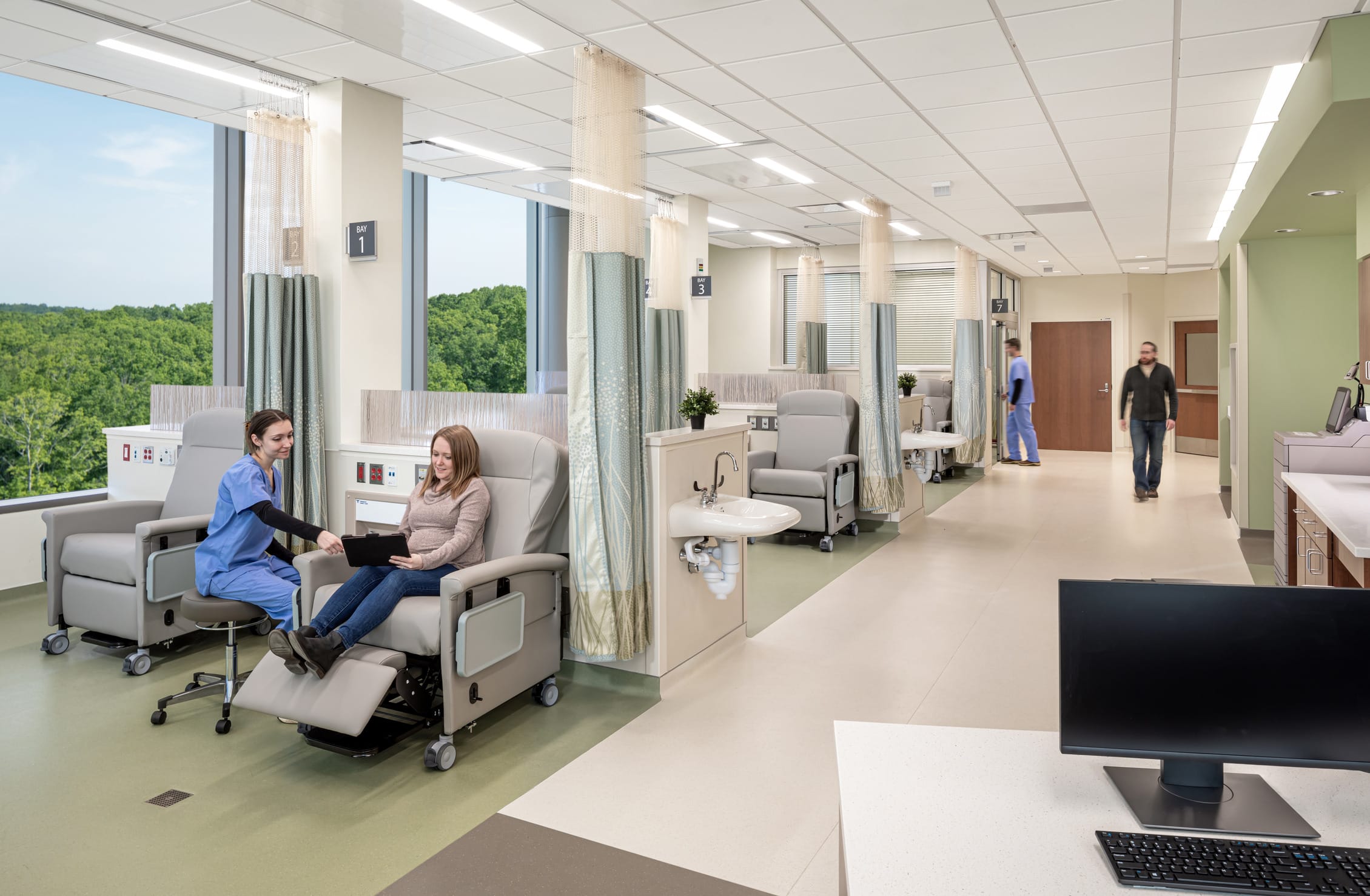 UNC Hospitals Hillsborough bright and modern treatment area with semi-private patient spaces separated by curtains. The room features reclining medical chairs with armrests, green flooring, and soft neutral-colored walls. Large windows allow natural light to enter, offering a view of the outdoors. A patient is receiving treatment in a chair, accompanied by a doctor in bright scrubs. This image showcases BSA's expertise in designing spaces that prioritize patient care.