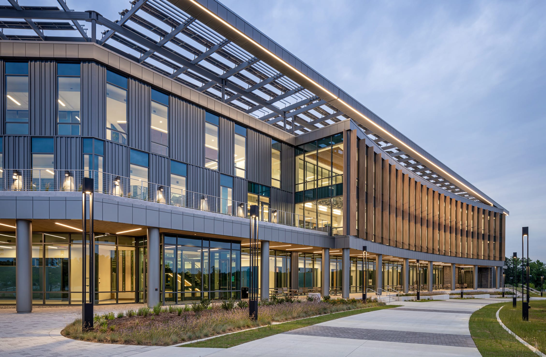 Angled exterior view of the Wake Tech Education and Innovation Center showcasing a dynamic façade with a mix of glass, metal, and vertical wood fins. The building features an expansive covered walkway supported by slender columns, large glass windows revealing interior activity, and a distinctive cantilevered roof with integrated lighting and structural framing overhead.