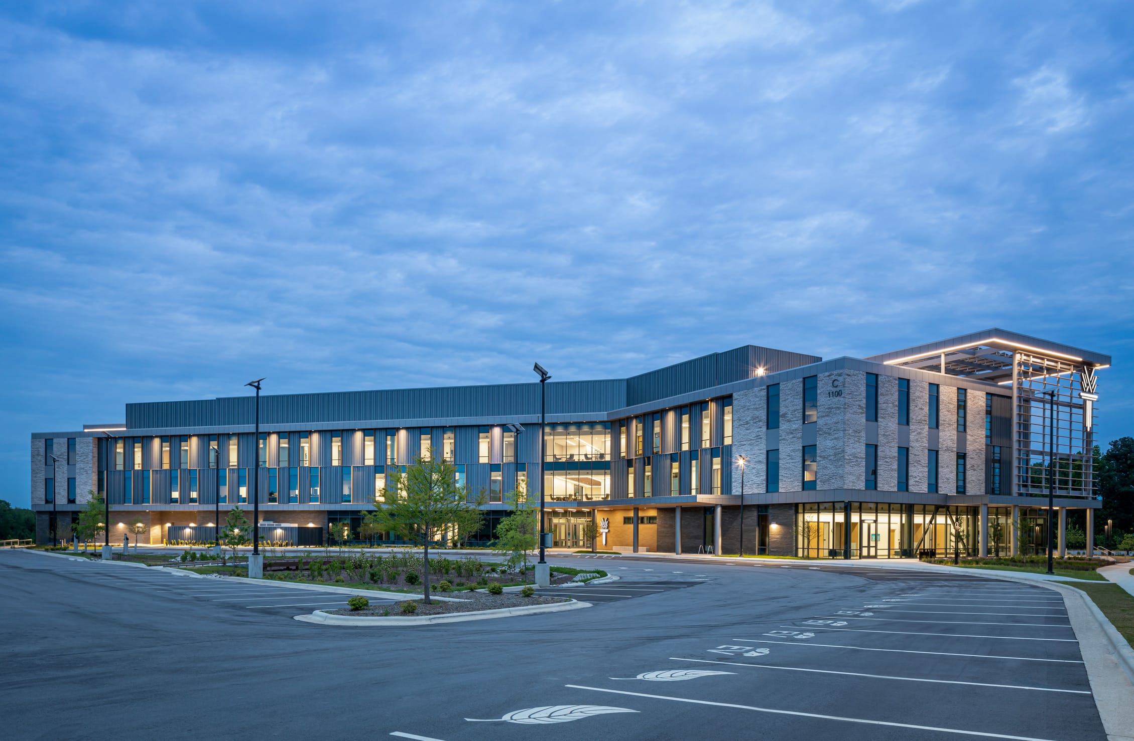 Exterior view of the Wake Tech Education and Innovation Center at dusk, designed with clean lines, expansive glass, and a prominent overhanging roof. The multi-story building features vertical fins, modern metal and brick finishes, and warm interior lighting that glows through the large windows, all set against a backdrop of open green space and a soft evening sky. This view shows an expansive parking lot in the foregorund.