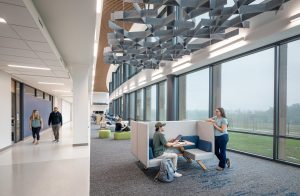 Interior view of a bright, modern student commons area at the Wake Tech Education and Innovation Center. The space features floor-to-ceiling windows with views of the green landscape outside. Overhead, an array of sculptural, geometric acoustic baffles hangs from the ceiling in dynamic, crisscrossed patterns, paired with linear pendant lighting. A slatted wood ceiling detail adds warmth and texture along the central spine of the space. Students are seen studying in upholstered booths, sitting on ottomans, and walking through the adjacent corridor, creating a lively and collaborative atmosphere.