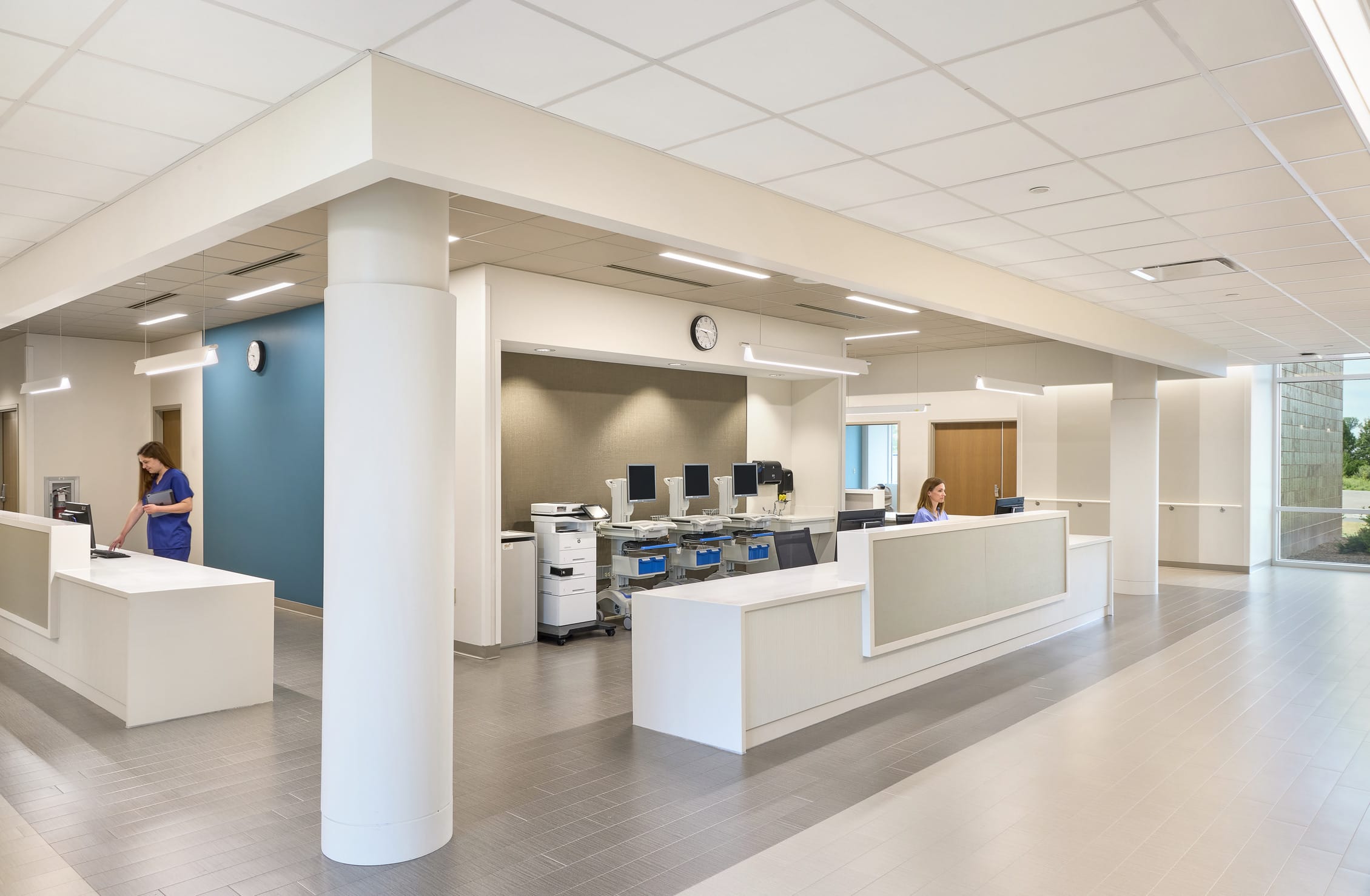 This image shows a modern nurses' station or administrative hub in what appears to be part of OrthoIndy Hospital. The space features a contemporary design with multiple white reception counters arranged in an open layout. Staff members in blue scrubs can be seen working at different stations. The area includes several workstations with multiple computer monitors, printers, and other office equipment in the central space. The design maintains the hospital's color scheme with white surfaces, blue accent walls, and neutral tones throughout. Linear lighting fixtures are integrated into the ceiling, providing even illumination across the space. The flooring continues the same pattern seen in the corridor image, with directional lines that help with wayfinding. Large structural columns support the ceiling in this open area, and there are windows visible on the far right that allow natural light to enter the space and provide views to the outside. This well-organized central hub appears designed to facilitate staff communication and efficient workflow in the healthcare environment.