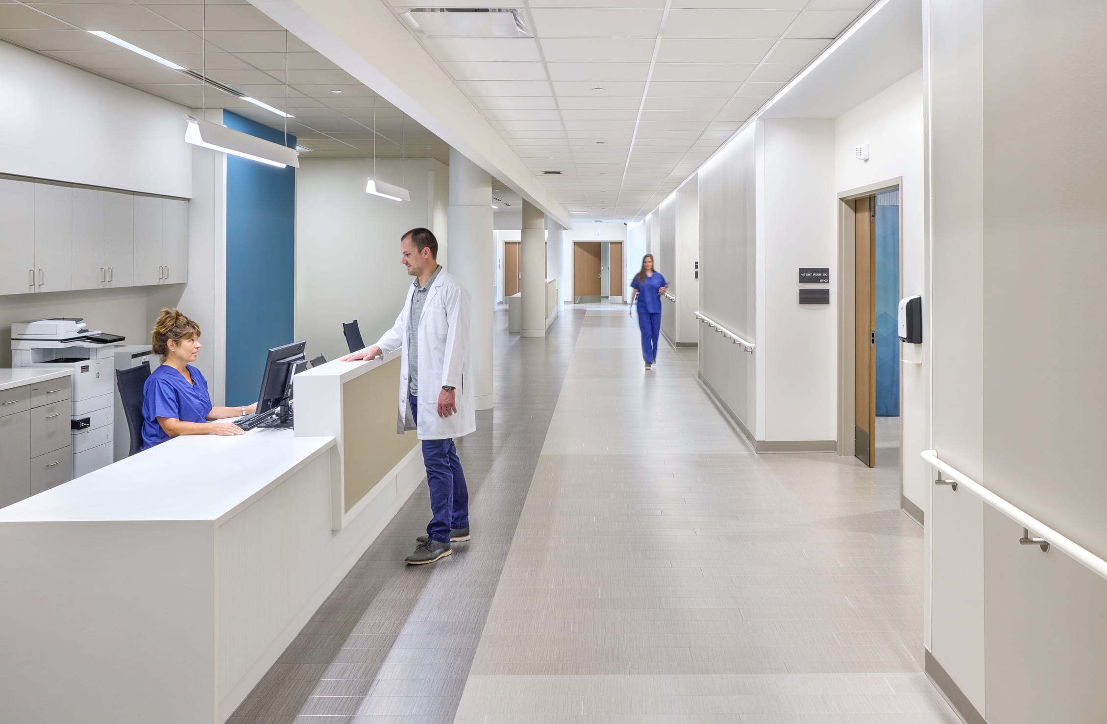 This image shows an interior corridor of what appears to be part of OrthoIndy Hospital. The space features a modern, clean design with a nurses' station or reception desk on the left where a staff member in blue scrubs is working at a computer. A healthcare professional in a white coat is standing at the counter conversing with the staff member. The corridor stretches down with a minimalist design featuring white walls accented with blue color panels. The flooring has a subtle pattern with directional lines that help guide movement through the space. Modern pendant lighting hangs from the drop ceiling, and there are handrails along the wall for patient support. In the distance, another staff member in blue scrubs can be seen walking down the hallway. The entire space conveys a contemporary healthcare environment with attention to both functionality and aesthetic design, consistent with the modern approach seen in the exterior architecture of the facility.