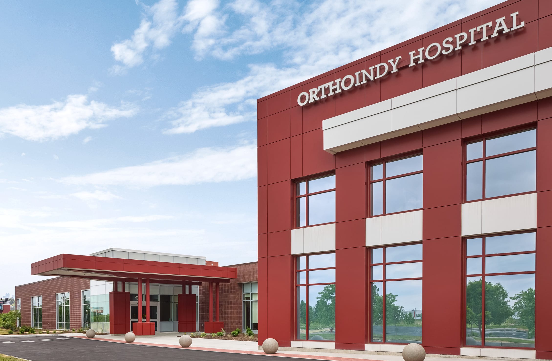 This image shows another exterior view of OrthoIndy Hospital, captured during daylight hours under a bright blue sky with scattered clouds. The building features a distinctive red and white façade with 