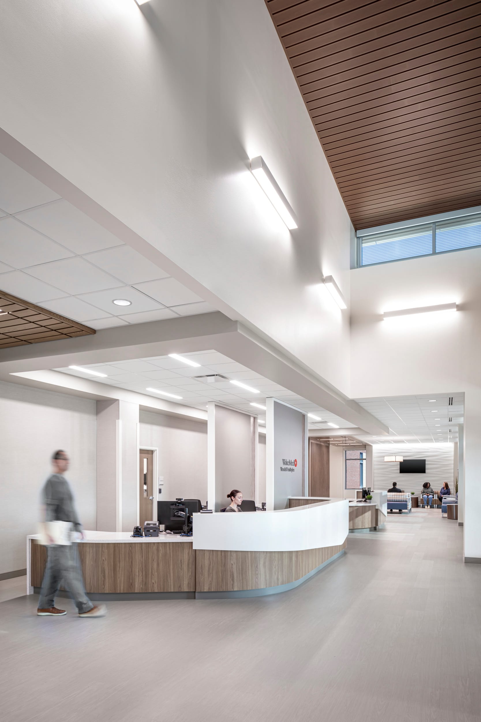 This image shows a modern healthcare facility, focusing on a reception area with a curved wood and white counter desk. A staff member is seated at the reception desk working at a computer terminal, while a person in gray clothing walks past carrying what looks like papers or documents. The space features a high ceiling with contemporary linear wall-mounted light fixtures and clerestory windows bringing in natural light. The ceiling combines white drop panels with wooden slat sections, creating visual warmth. In the background, a waiting area is visible with several people seated, and there appears to be signage on the wall that might indicate the name of the facility. The overall design maintains the clean, bright aesthetic with neutral tones, combining functionality with a welcoming atmosphere typical of modern healthcare environments.