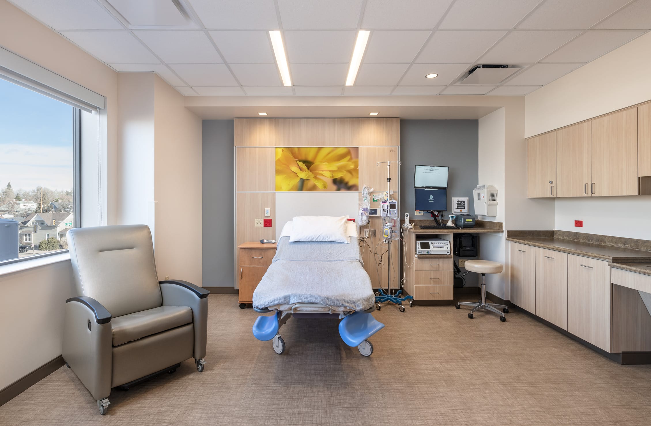 This image shows another patient room in the Cheyenne Regional Medical Center's labor and delivery unit, this one featuring a more spacious layout and expansive window views. The room includes a hospital bed with light blue linens and stirrup attachments, positioned in front of a headwall featuring a yellow floral image that continues the nature-inspired design theme seen throughout the unit. To the left is a comfortable gray mobile recliner chair for patient or family use. The large window offers a scenic view of the surrounding Cheyenne neighborhood with trees and houses visible in the distance. The room includes built-in cabinetry and counter space on the right, a small workstation with stool, and necessary medical equipment including monitors and IV poles. The medical headwall contains integrated outlets and connections while maintaining a residential feel with its light wood finishes. The room's design provides both clinical functionality for childbirth and a tranquil, healing environment with natural light and views.