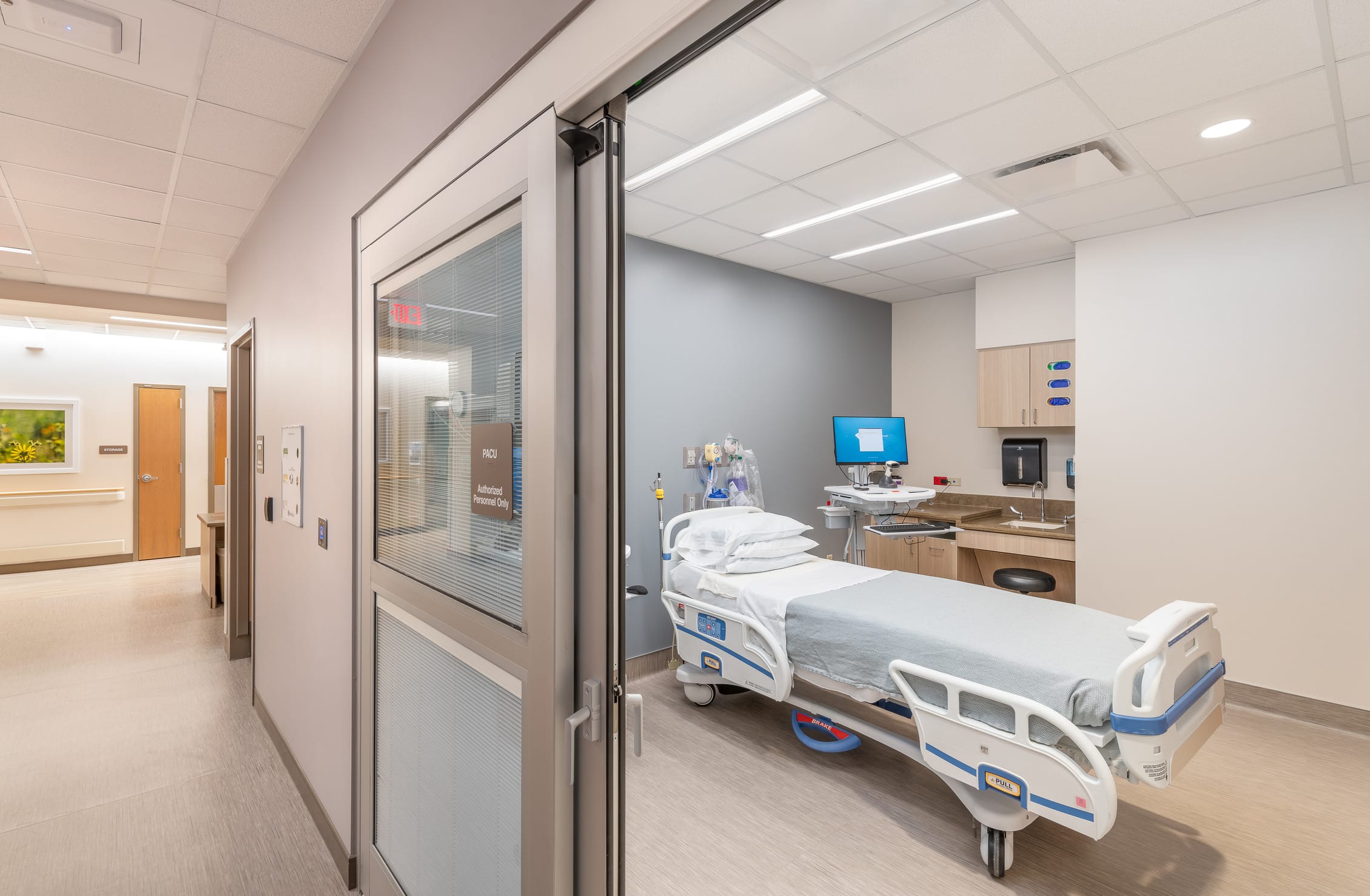 This image shows a modern hospital patient room in what appears to be the Cheyenne Regional Medical Center. The view is taken from the hallway, looking through a sliding glass door with integrated blinds that's partially open. The spacious room features a standard hospital bed with white linens and an electronic control panel visible on its side. The room has a light blue accent wall and is equipped with medical monitoring equipment, a computer workstation on the right side, and storage cabinets. Medical supplies and equipment are neatly arranged, and the room has recessed lighting in a dropped ceiling. The hallway visible on the left appears bright and well-maintained with what looks like artwork on the wall, creating a welcoming clinical environment that balances medical functionality with patient comfort.