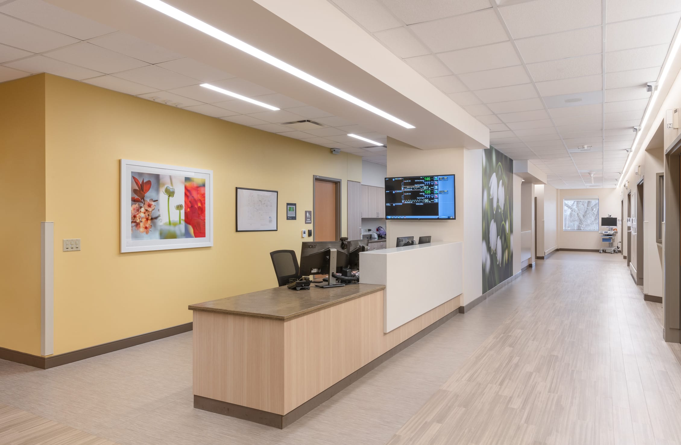 This image shows another nursing station in the Cheyenne Regional Medical Center's labor and delivery unit. The station features a modern design with light wood cabinetry, white countertops, and gray work surfaces. A large wall-mounted monitor displays patient monitoring data. The station is positioned along a corridor with patient rooms, creating a central hub for staff to monitor and respond to patients' needs. The warm yellow accent wall adds a cheerful element to the space and displays framed nature photography with red and green floral imagery, continuing the botanical theme seen throughout the unit. Beyond the desk, the corridor extends with the same light wood-look flooring in a herringbone pattern, and another floral wall mural is visible in the distance. The design creates a professional yet welcoming medical environment that balances clinical functionality with aesthetic warmth.