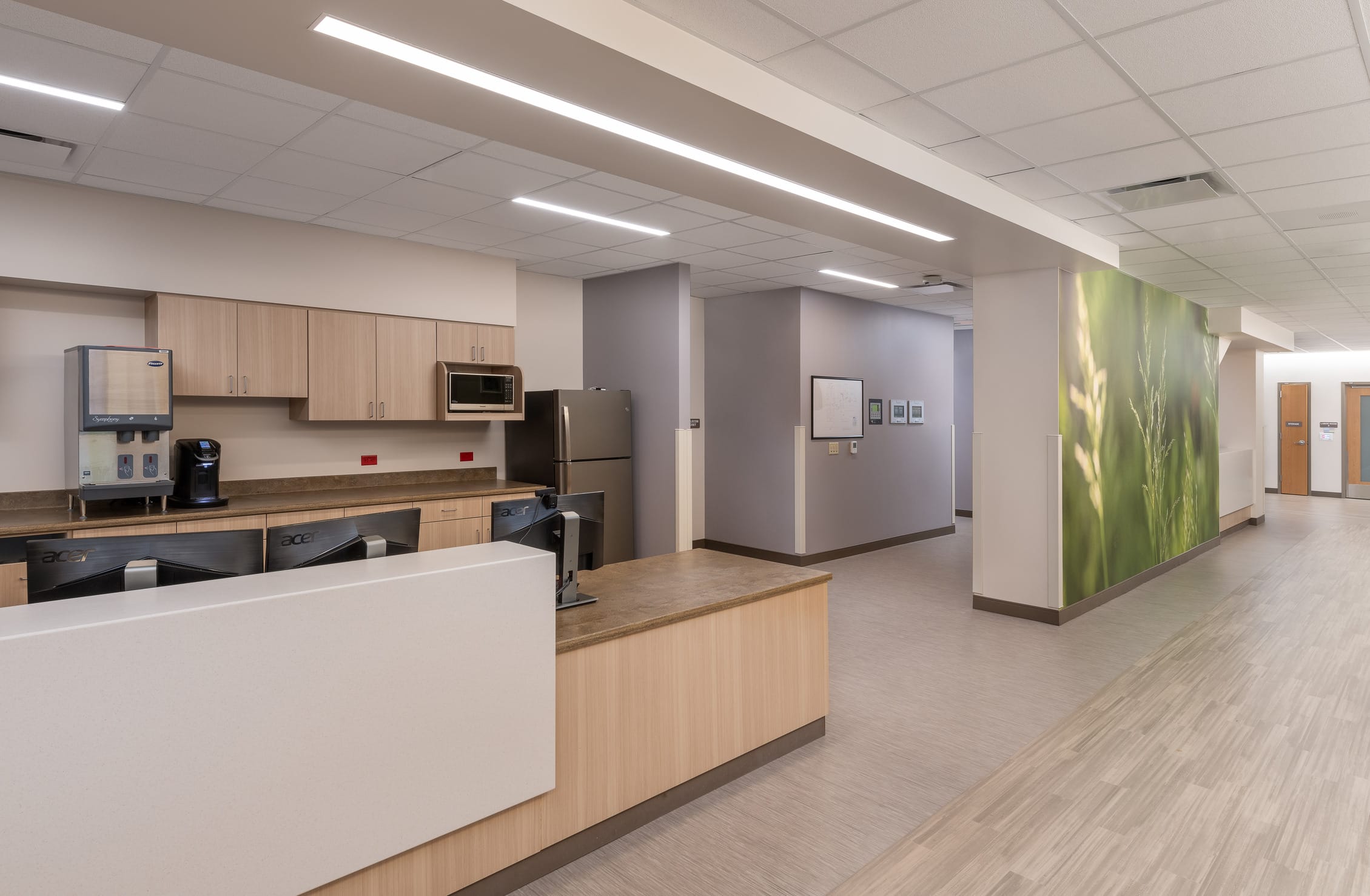 This image shows a modern nurses' station and staff area within the Cheyenne Regional Medical Center. The space features a reception counter with light wood cabinetry and brown countertops, behind which is a well-equipped break area including a coffee maker, water dispenser, microwave, and refrigerator. Computer workstations with Acer monitors are visible for staff use. The area has a clean, contemporary design with soft gray walls, light wood-look flooring, and recessed lighting in a white ceiling grid. To the right of the image is a hallway with a striking nature-themed wall mural of green grass or wheat, adding a calming biophilic element to the clinical environment. Small framed artwork can be seen on the opposite wall, and the open layout allows for easy staff movement and communication while maintaining a professional healthcare atmosphere.