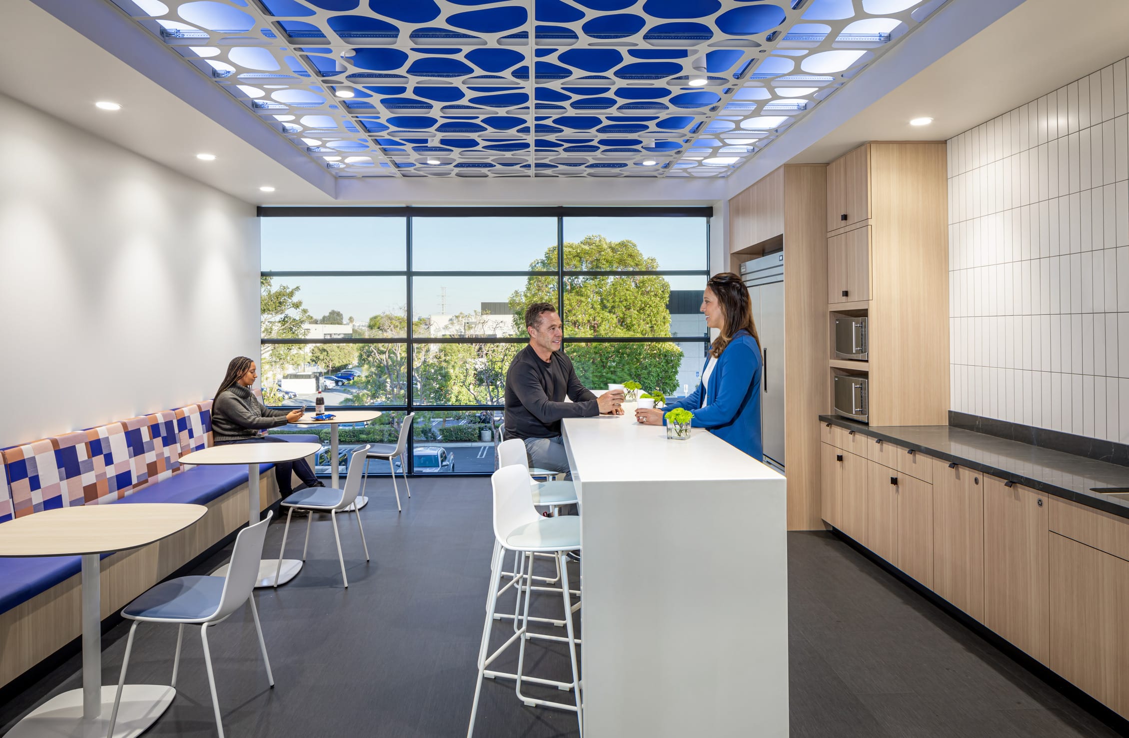 Bright break room in the BSA-designed histology facility featuring the signature cellular-patterned ceiling with blue backlighting that unifies the design throughout the space. Two colleagues converse at a central white island counter while another works independently at a café table. Floor-to-ceiling windows flood the room with natural light and provide verdant views of surrounding trees. The functional kitchenette includes light wood cabinetry, integrated appliances, and a colorful patterned banquette seating area with round tables, creating a comfortable space for staff to relax and collaborate away from laboratory settings.
