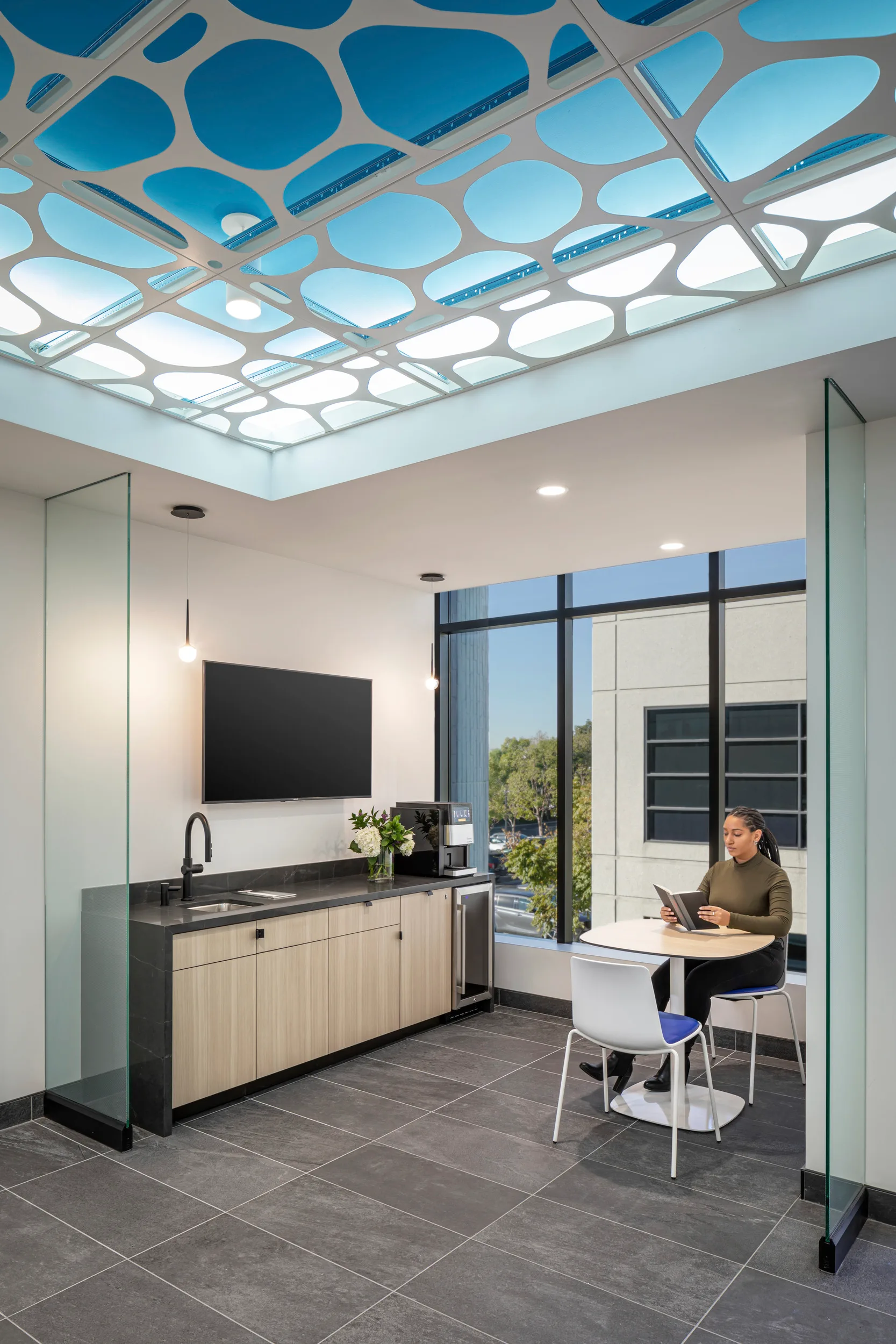 Intimate breakout space in the BSA-designed histology facility featuring the signature cellular-patterned ceiling with blue and white backlit panels. A staff member reviews material on a tablet at a round table with contemporary white and blue chairs, beside floor-to-ceiling windows offering exterior views. The compact kitchenette includes light wood cabinetry, dark stone countertops, integrated sink, and a wall-mounted screen for presentations or media viewing. Minimalist pendant lighting and glass partition walls create an open feeling within the compact space, maintaining the facility's cohesive modern aesthetic with gray tile flooring.