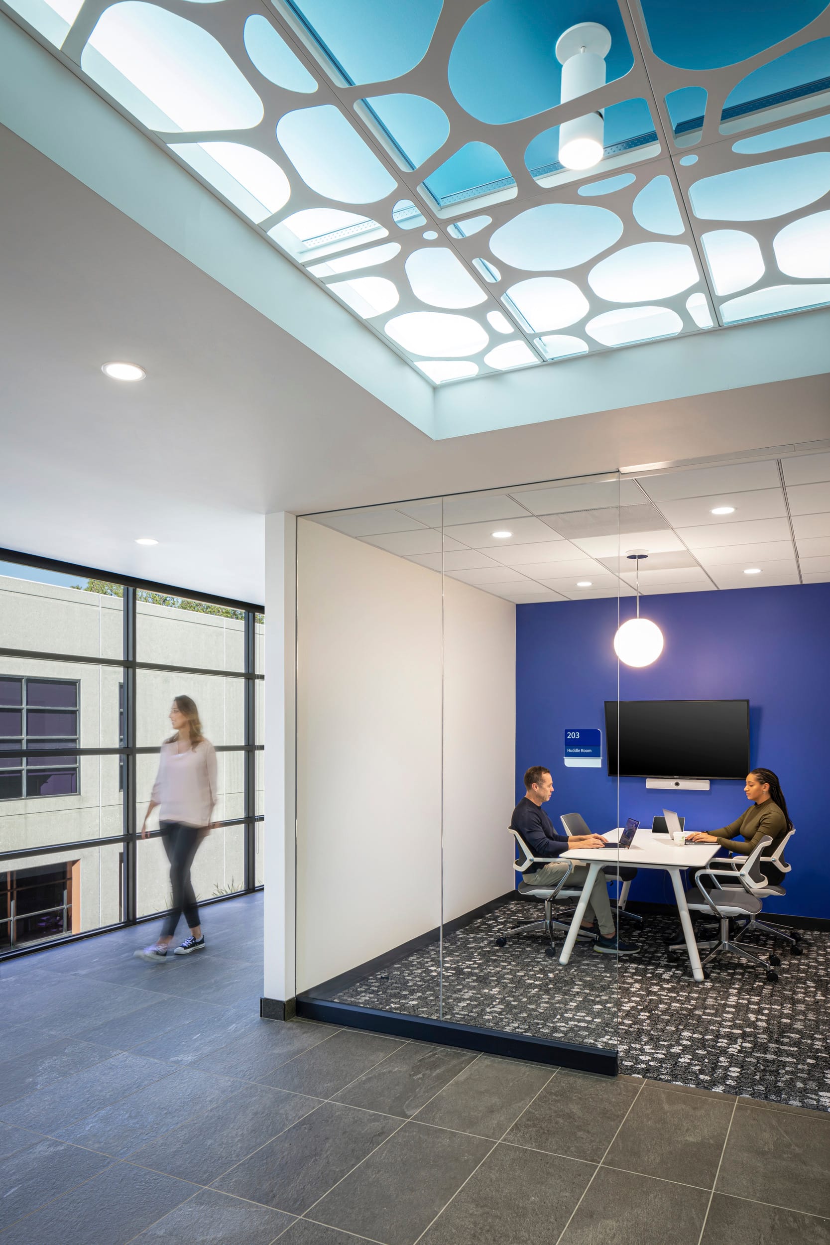 Conference room in the BSA-designed histology facility featuring glass walls for transparency and the distinctive cellular-patterned ceiling with blue backlighting. Two colleagues work collaboratively at a white meeting table with laptops, against a vibrant royal blue accent wall with wall-mounted display screen. The room is identified as '203' with appropriate signage and features a contemporary globe pendant light. Outside, a staff member walks along the corridor with large windows providing natural light and exterior views, maintaining the facility's modern aesthetic with consistent gray tile flooring and patterned carpeting within the meeting space.
