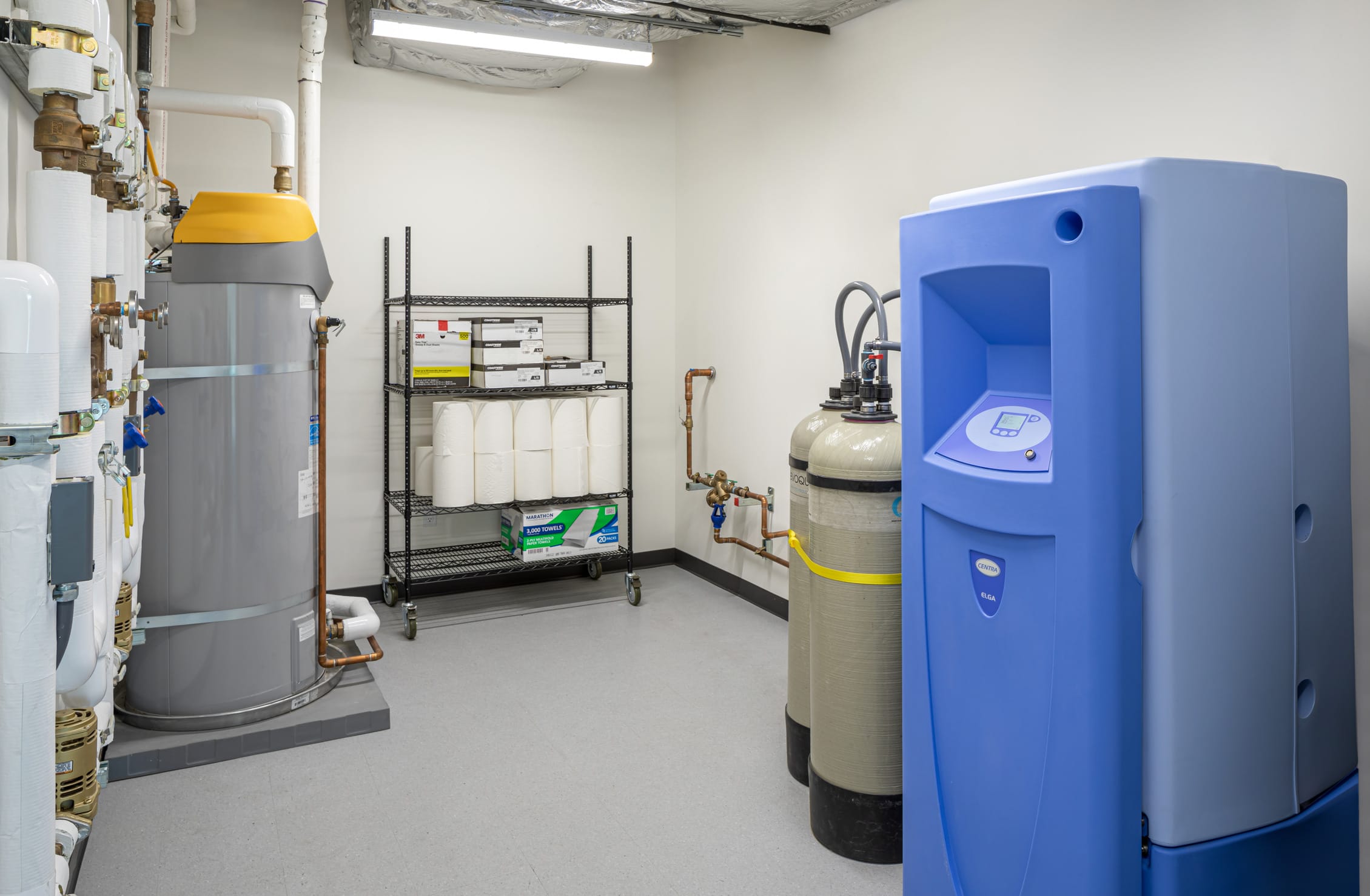 Interior of a histology lab utility room featuring water treatment equipment including a gray water heater with yellow top on the left, a blue water purification system on the right, and two beige filtration tanks in the center. The room contains plumbing infrastructure with copper pipes and valves mounted on white walls, and a metal storage rack holding paper supplies and boxed materials. The clean, clinical space has a white floor and fluorescent lighting overhead.
