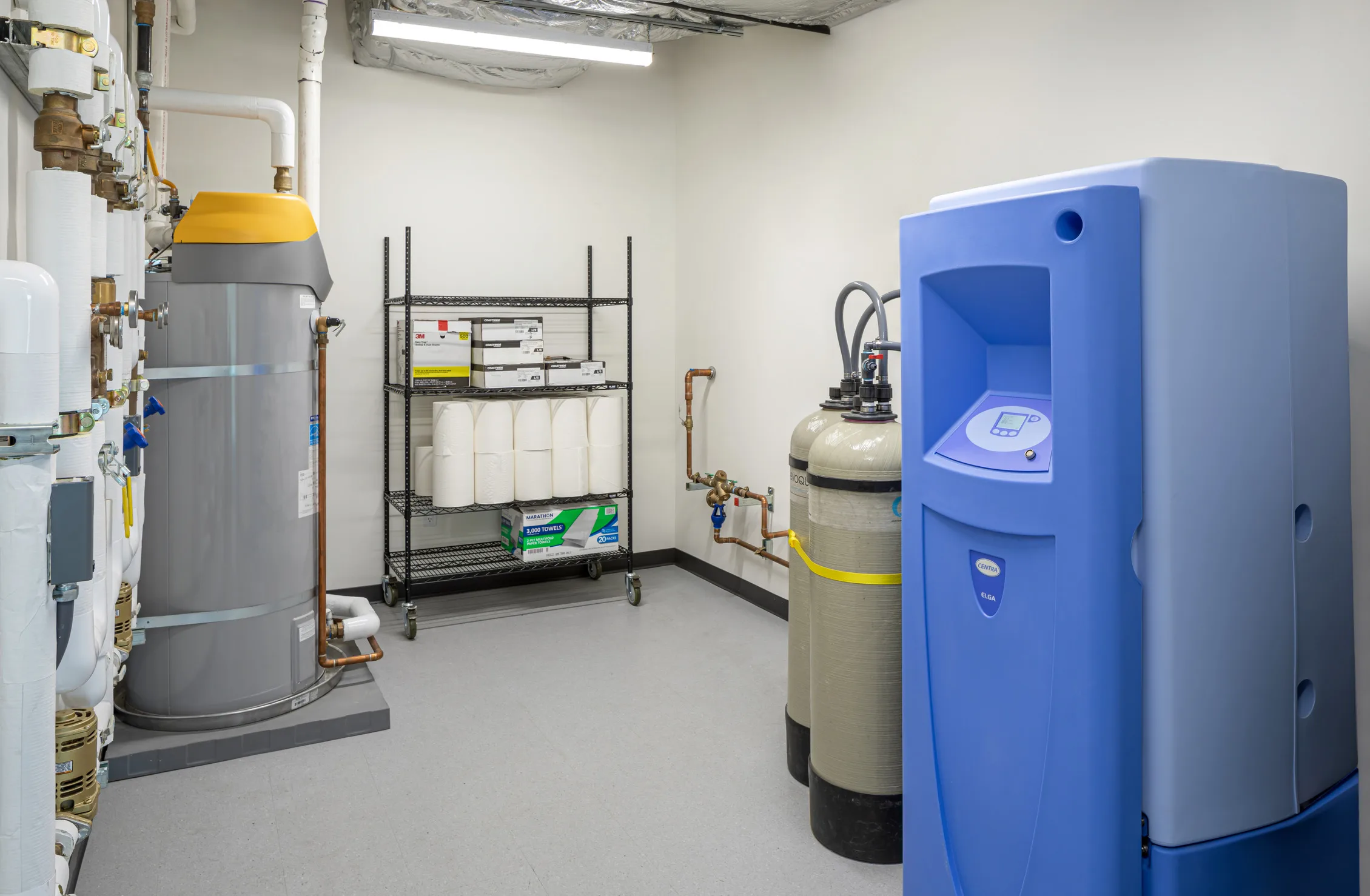 Interior of a histology lab utility room featuring water treatment equipment including a gray water heater with yellow top on the left, a blue water purification system on the right, and two beige filtration tanks in the center. The room contains plumbing infrastructure with copper pipes and valves mounted on white walls, and a metal storage rack holding paper supplies and boxed materials. The clean, clinical space has a white floor and fluorescent lighting overhead.