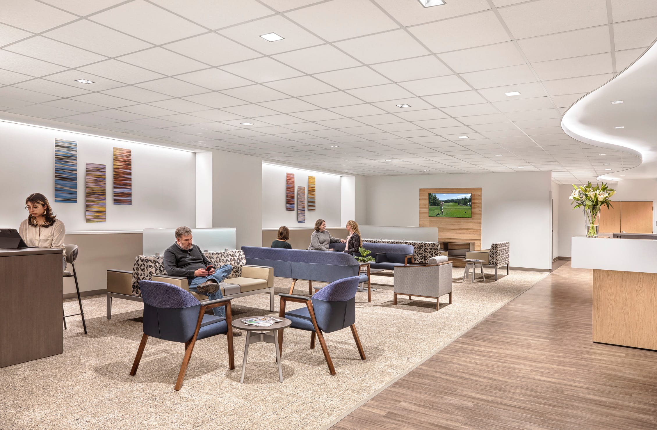 The image shows a modern healthcare waiting area inside Jacksonville Memorial Health. The spacious lounge features a contemporary design with comfortable seating arrangements including blue armchairs with wooden legs, patterned sofas, and various seating clusters throughout the space. Several people are present - a woman at what appears to be a reception desk, a man looking at reading material, and two women conversing in the background. The interior has a calming aesthetic with white drop ceiling tiles, recessed lighting, colorful abstract art panels on the walls, a wall-mounted screen displaying a golf course scene, and natural wood accents. A reception counter is visible to the right with fresh white lilies in a vase. The flooring transitions from light carpet in the seating area to wood-look flooring near the entrance, creating a welcoming and professional healthcare environment.