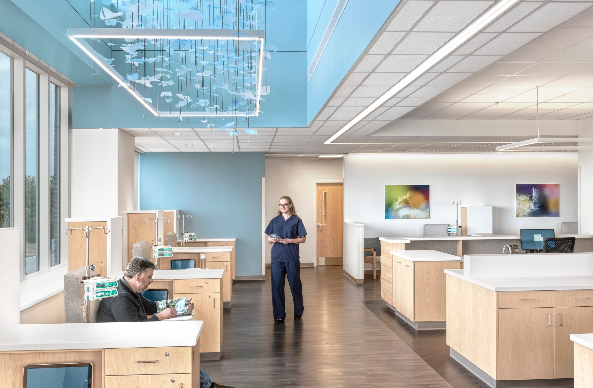 This image shows an infusion center within Jacksonville Memorial Health. The space features a modern medical treatment area with individual infusion stations equipped with light wood cabinetry and white countertops. A healthcare worker wearing navy blue scrubs stands in the center of the room, while a patient sits in one of the treatment areas on the left side. The design incorporates calming blue accent walls complemented by large windows that allow natural light. A striking decorative installation hangs from a high ceiling area with suspended white elements creating a soothing visual effect. The infusion room includes medical equipment such as IV poles, comfortable treatment chairs, and workstations for staff. Nature-inspired artwork on the walls (featuring what appears to be dandelion imagery) adds to the healing atmosphere. The overall design balances clinical functionality with elements intended to create a comfortable environment for patients receiving treatment.