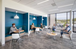This image highlights a thoughtfully designed waiting area at the Dearborn Cancer Center by BSA, offering diverse seating arrangements that accommodate different needs and preferences of patients and visitors. Along the left wall, three semi-private banquette alcoves with vibrant teal blue backing create intimate spaces for focus or quiet conversation, each illuminated by modern pendant lighting and featuring round wooden tables. The first alcove shows a young man working on a laptop, while the middle alcove hosts two women in conversation. The right side of the space offers a more traditional lounge setting with comfortable armchairs arranged around circular tables, where two people are engaged with reading materials. Floor-to-ceiling windows flood the space with natural light and provide expansive views of the surrounding landscape and parking area. The muted gray carpeting, white ceiling with recessed lighting, and natural wood accents maintain the facility's calming color palette. This versatile waiting environment demonstrates BSA's understanding of how different patients and families might need varying levels of privacy and comfort during cancer treatment visits.