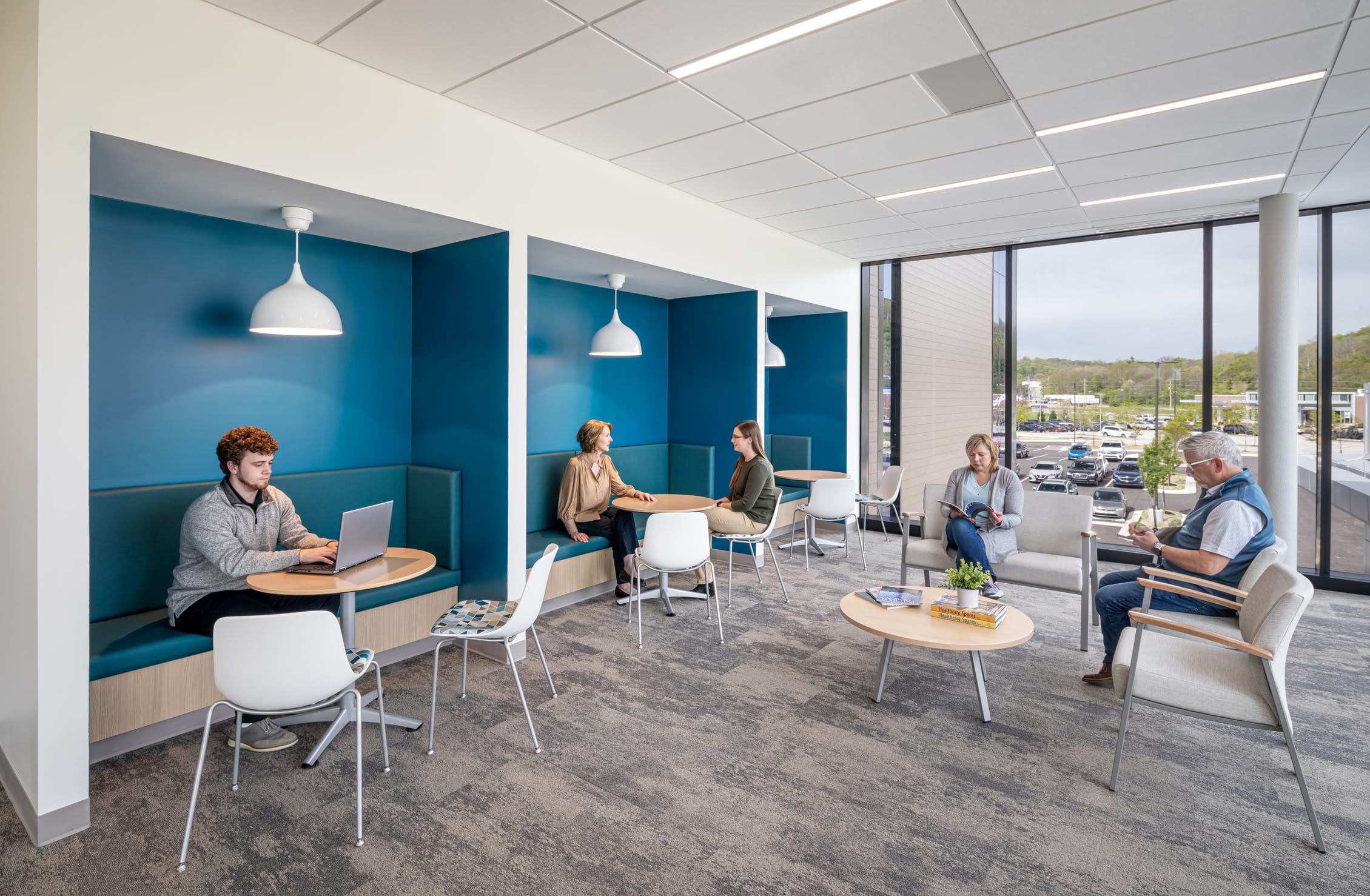 This image highlights a thoughtfully designed waiting area at the Dearborn Cancer Center by BSA, offering diverse seating arrangements that accommodate different needs and preferences of patients and visitors. Along the left wall, three semi-private banquette alcoves with vibrant teal blue backing create intimate spaces for focus or quiet conversation, each illuminated by modern pendant lighting and featuring round wooden tables. The first alcove shows a young man working on a laptop, while the middle alcove hosts two women in conversation. The right side of the space offers a more traditional lounge setting with comfortable armchairs arranged around circular tables, where two people are engaged with reading materials. Floor-to-ceiling windows flood the space with natural light and provide expansive views of the surrounding landscape and parking area. The muted gray carpeting, white ceiling with recessed lighting, and natural wood accents maintain the facility's calming color palette. This versatile waiting environment demonstrates BSA's understanding of how different patients and families might need varying levels of privacy and comfort during cancer treatment visits.