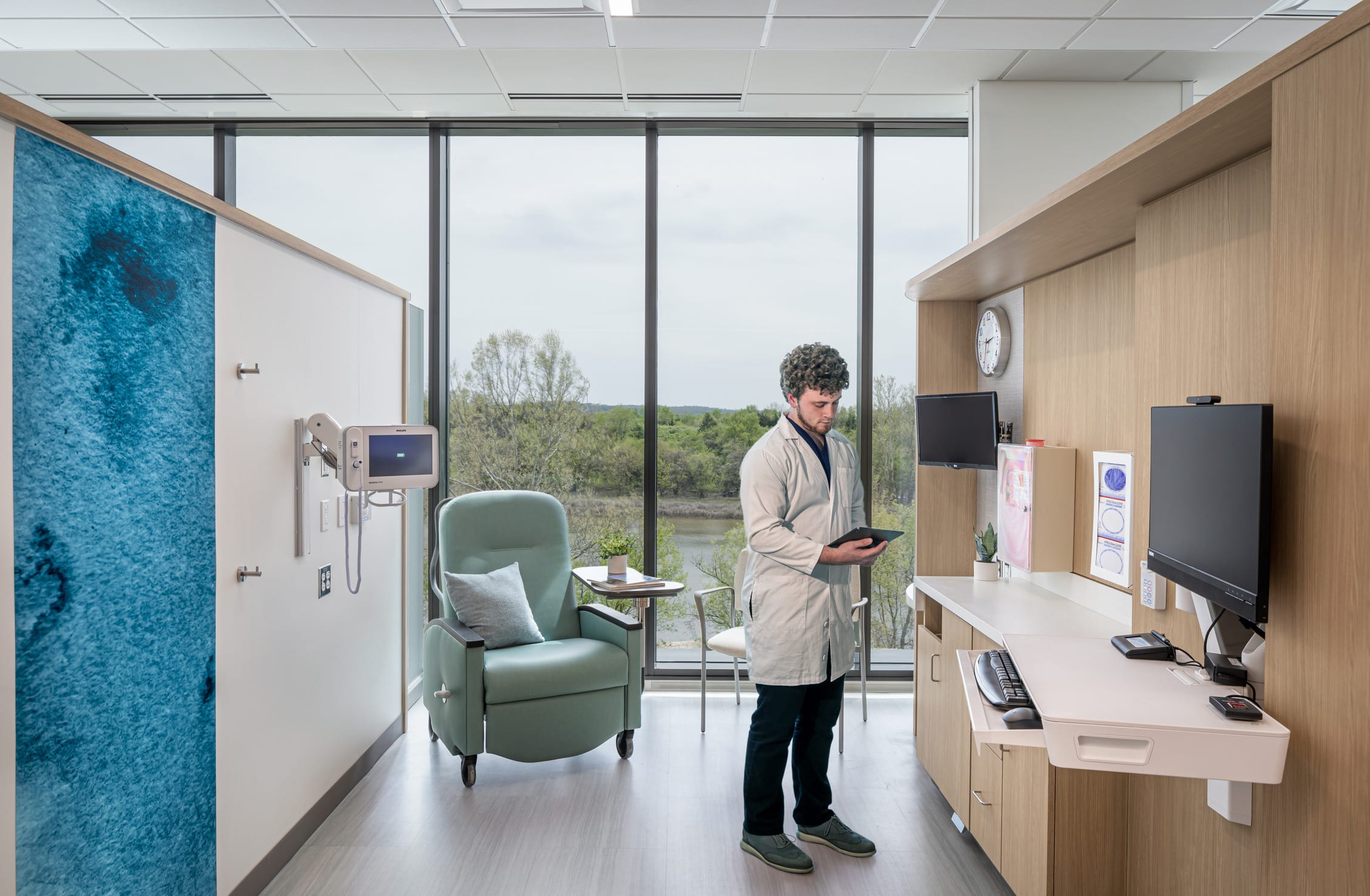 This image showcases a patient treatment room at the Dearborn Cancer Center by BSA, designed to balance clinical functionality with comfort and connection to nature. The room features floor-to-ceiling windows providing expansive views of natural landscaping and water, creating a calming backdrop for treatment. A healthcare provider in a light lab coat reviews information on a tablet near the built-in workstation with dual monitors and medical reference materials. The patient area includes a comfortable mint-green reclining treatment chair with a pillow, positioned for both treatment access and enjoyment of the outdoor views. Clinical necessities are thoughtfully integrated, including wall-mounted medical equipment and monitors, while maintaining a non-institutional atmosphere. The design incorporates BSA's signature blue glass panel element at the entrance, warm wood-look cabinetry for medical supplies and equipment, and light-colored flooring, all working together to create a healing environment that supports both efficient care delivery and patient wellbeing during what can be stressful treatment sessions.