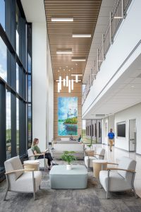 This image showcases the modern and welcoming interior of the Dearborn Cancer Center designed by BSA. The spacious lobby features floor-to-ceiling windows along the left side, flooding the space with natural light and offering views of the landscaped grounds outside. In the foreground, a comfortable waiting area includes light-colored armchairs arranged around a wooden circular table on a textured carpet.