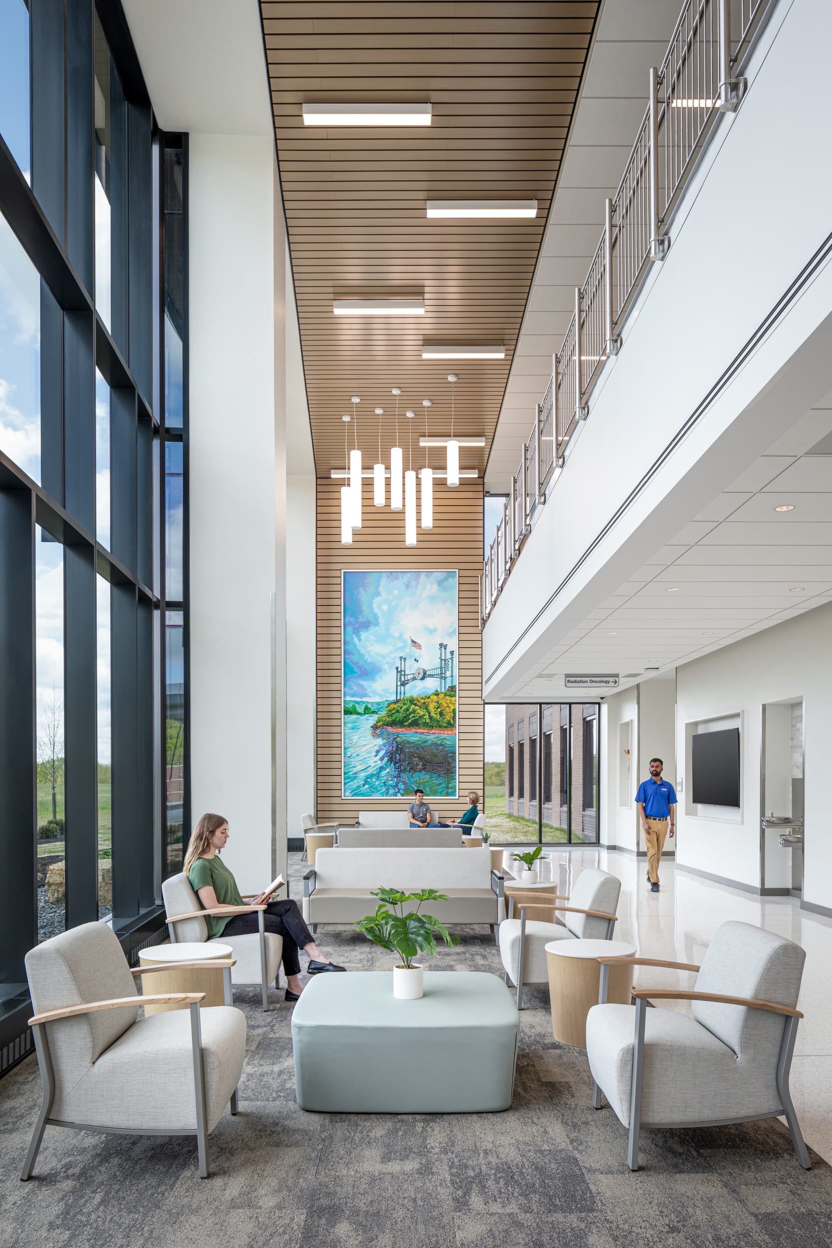 This image showcases the modern and welcoming interior of the Dearborn Cancer Center designed by BSA. The spacious lobby features floor-to-ceiling windows along the left side, flooding the space with natural light and offering views of the landscaped grounds outside. In the foreground, a comfortable waiting area includes light-colored armchairs arranged around a wooden circular table on a textured carpet.