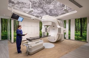 This image showcases a state-of-the-art radiation treatment room at the Dearborn Cancer Center designed by BSA, where advanced medical technology meets thoughtful, patient-centered design. The space features a sophisticated TrueBeam radiation therapy system in sleek white, positioned prominently alongside a specialized treatment table. A healthcare professional in navy blue scrubs stands attentively at the control panel. What distinguishes this clinical space is the biophilic design approach with floor-to-ceiling forest murals wrapping around the walls, bringing elements of nature indoors to create a calming environment for patients during treatment. The ceiling features a decorative perforated metal panel with subtle lighting fixtures, adding visual interest while maintaining the room's soothing aesthetic. Built-in white cabinetry provides necessary storage while maintaining the clean, uncluttered appearance of the space. The warm wood-look flooring enhances the natural theme and contributes to the room's less institutional, more comforting atmosphere.