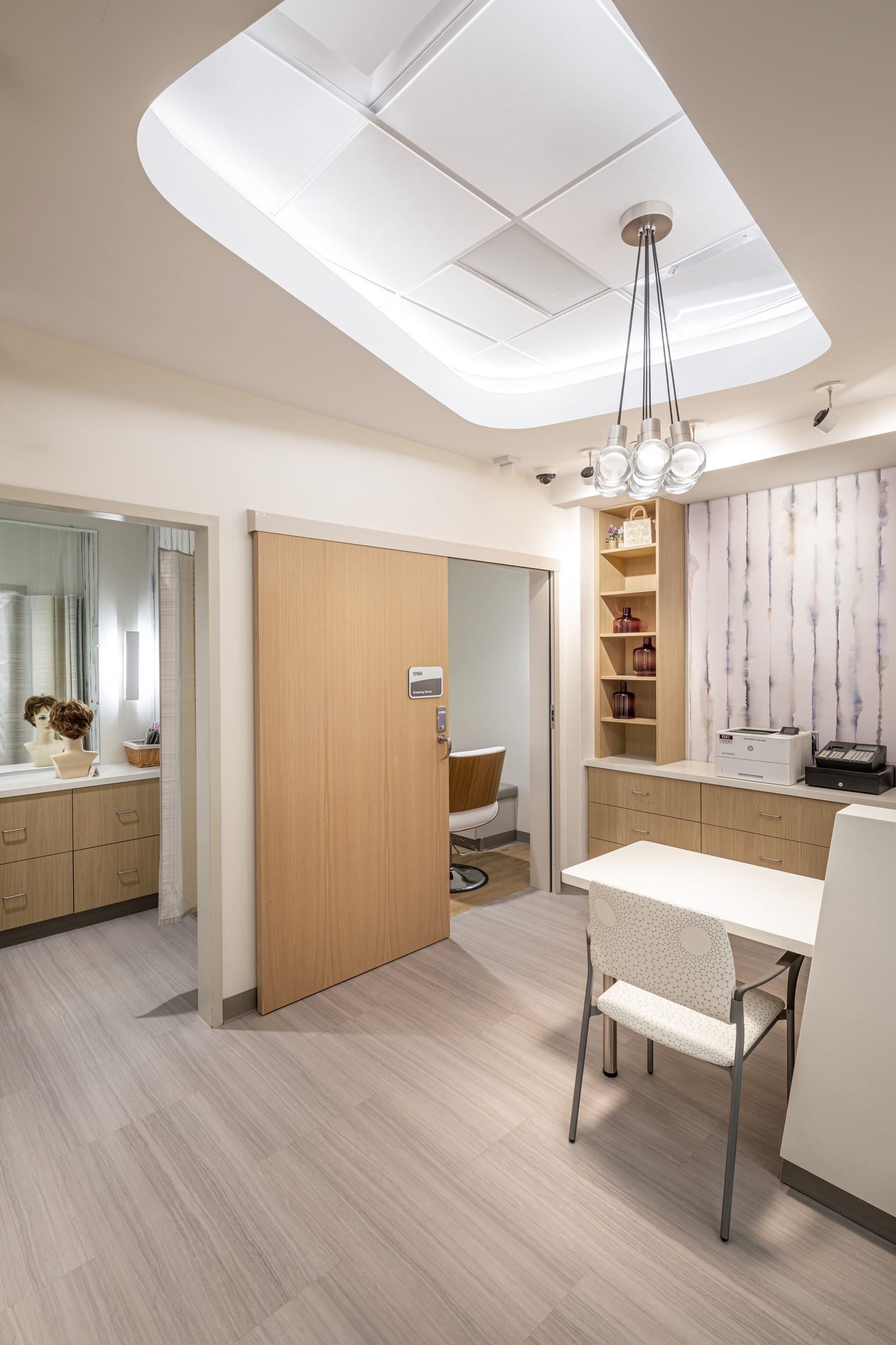 This thoughtfully designed space at the Dearborn Cancer Center by BSA reflects a warm, welcoming, and dignified environment for patients. Soft wood tones, natural textures, and layered lighting—especially the sculptural pendant and cove ceiling detail—enhance a sense of calm and comfort. The inclusion of a private consultation or wig-fitting area acknowledges the sensitive needs of cancer patients, while open shelving and a modern work surface provide functionality for staff. The overall aesthetic promotes healing through its balance of privacy, beauty, and efficiency.