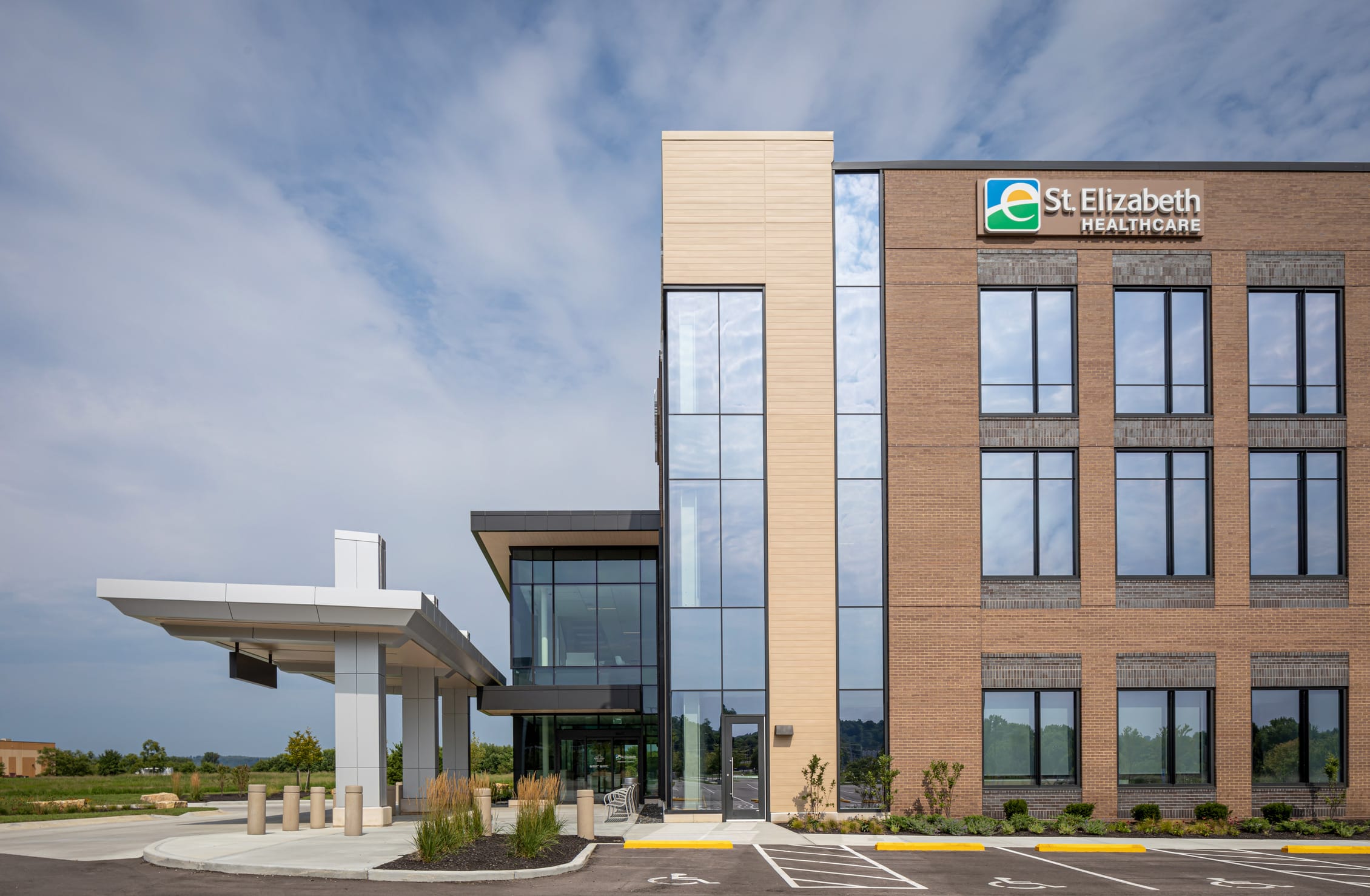 This view of the Dearborn Cancer Center emphasizes its architectural clarity and strong material palette, where warm brick, light wood tones, and expansive glass convey a sense of trust and transparency. The juxtaposition of solid and transparent elements highlights both permanence and openness—key values in a healthcare setting. The covered drop-off canopy ensures patient comfort and accessibility, while thoughtfully placed landscaping softens the urban edge and contributes to a welcoming, non-institutional atmosphere.