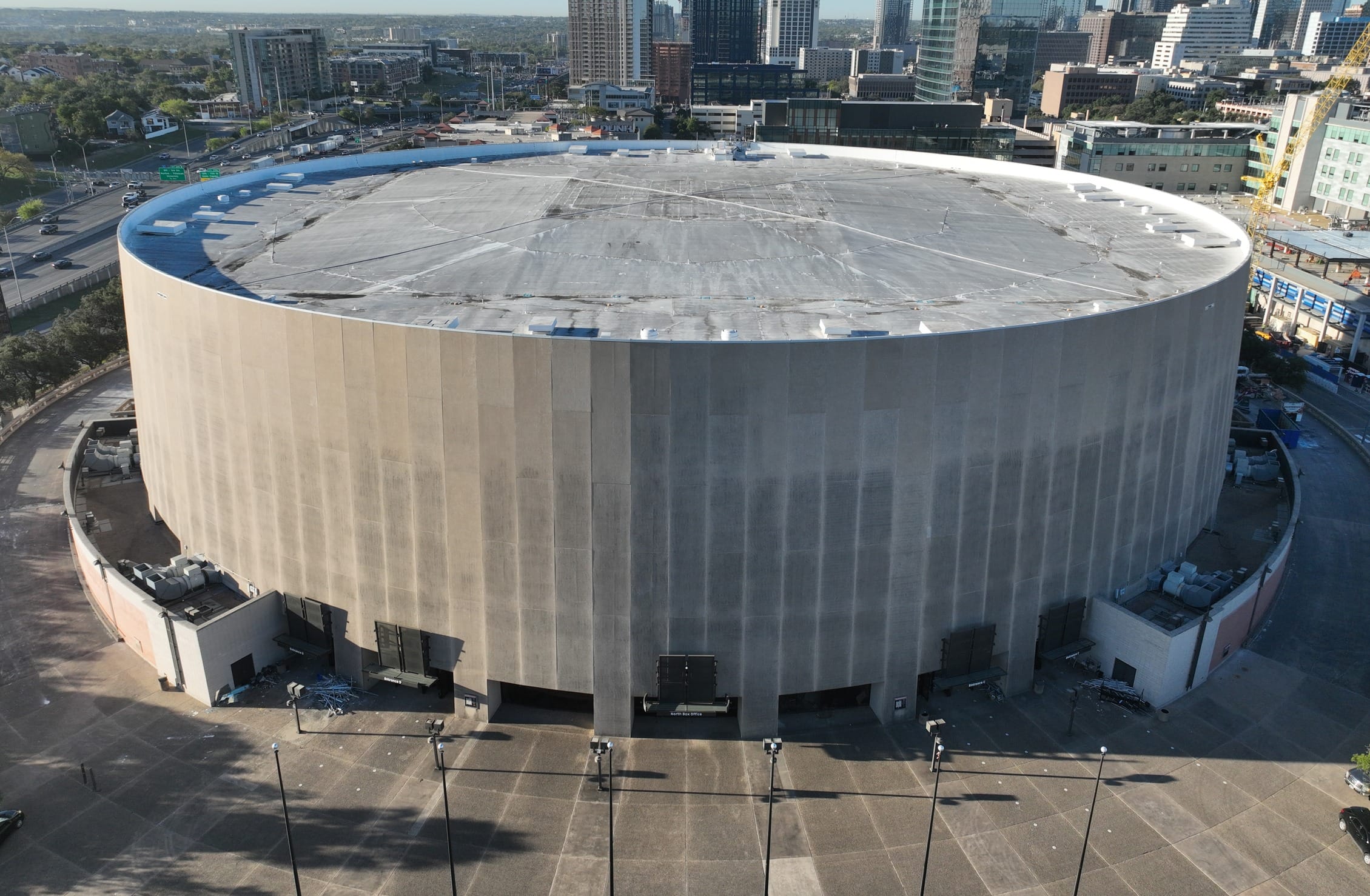 Aerial view of the Frank Erwin Center in Austin, Texas, before demolition, showing the iconic drum-shaped arena with its distinctive circular white roof. The facility is positioned alongside a major highway with the Austin downtown skyline rising prominently in the background. New construction is visible adjacent to the arena, with development cranes and buildings under construction to the right. The image captures the urban context of the venue with surrounding parking areas, roadways, and the lush green landscape of Austin extending toward the horizon.