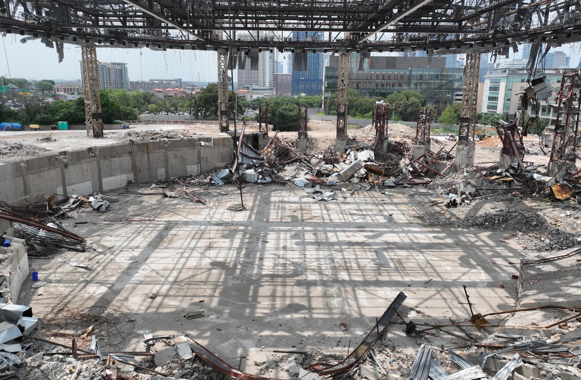 Interior view of the Frank Erwin Center demolition in Austin, showing the concrete floor of the former arena surrounded by debris and structural steel. The remaining roof framework casts geometric shadows across the floor while supporting columns stand partially intact. Through the skeletal structure, the Austin skyline is visible in the background, with modern high-rises and greenery contrasting against the demolition site.