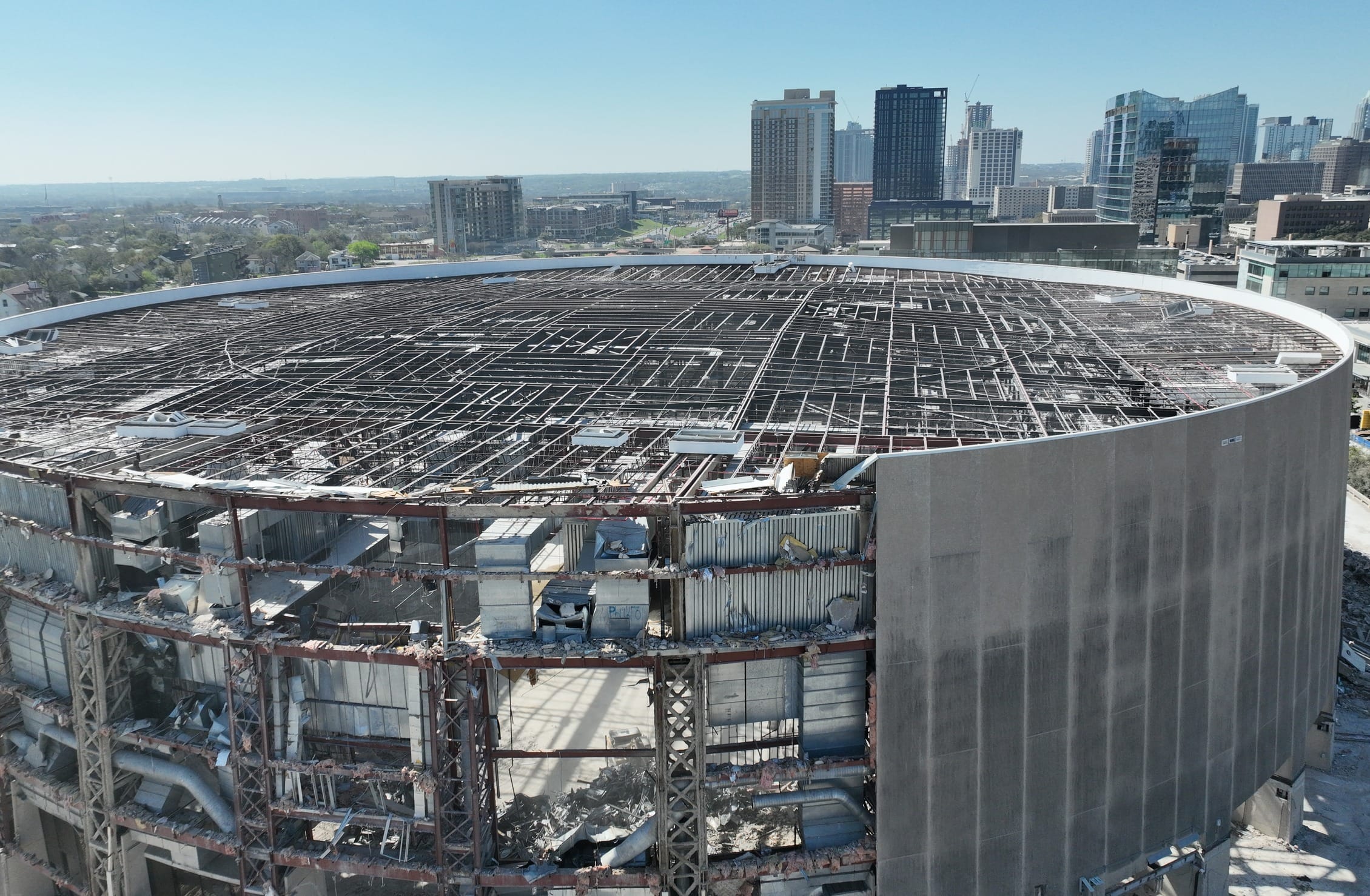 Aerial view of the partial demolition of the Frank Erwin Center in Austin, Texas, showing the circular steel framework of the arena being dismantled. Construction equipment surrounds the site as the once-iconic venue is being torn down. The downtown Austin skyline with high-rise buildings is visible in the background, along with busy roadways, parking lots, and green spaces stretching into the distance.