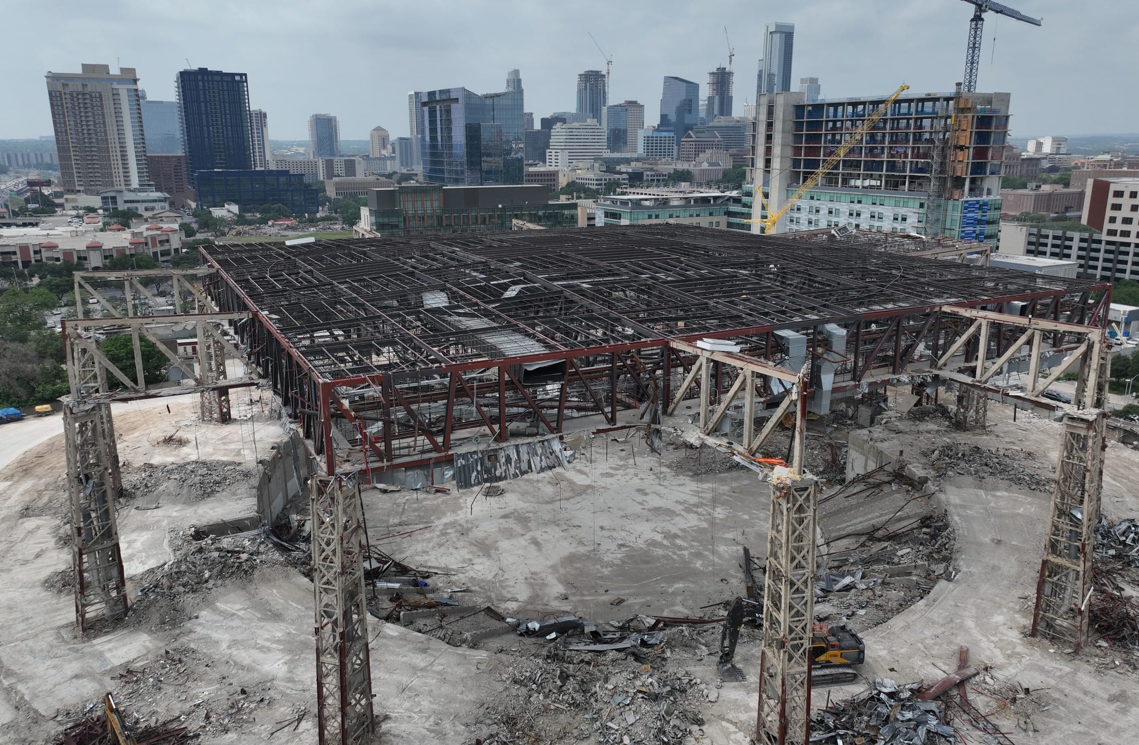 Aerial view of the partial demolition of the Frank Erwin Center in Austin, Texas, showing the circular steel framework of the arena being dismantled. Construction equipment surrounds the site as the once-iconic venue is being torn down. The downtown Austin skyline with high-rise buildings is visible in the background.