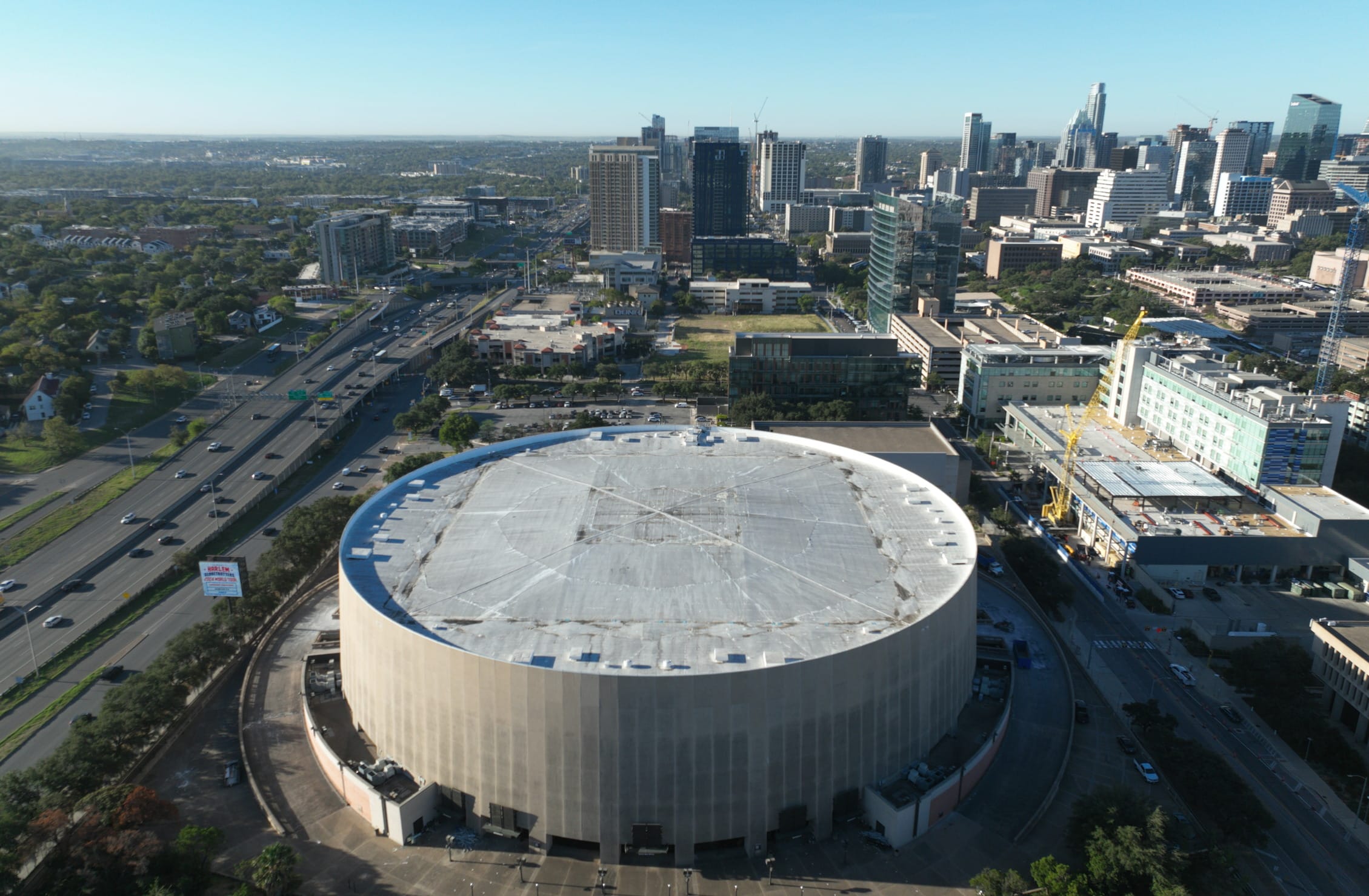 Aerial view of the Frank Erwin Center in Austin, Texas, before demolition, showing the iconic drum-shaped arena with its distinctive circular white roof. The facility is positioned alongside a major highway with the Austin downtown skyline rising prominently in the background. New construction is visible adjacent to the arena, with development cranes and buildings under construction to the right. The image captures the urban context of the venue with surrounding parking areas, roadways, and the lush green landscape of Austin extending toward the horizon.