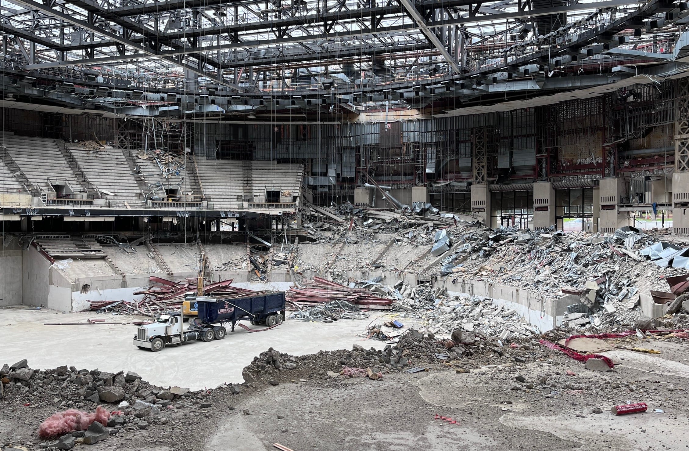 Interior demolition of the Frank Erwin Center arena floor, with a semi-trailer truck positioned amid piles of rubble and debris. Remnants of the lower bowl seating are visible in the background, while the upper steel framework and roof structure remain intact. The once-bustling event space is now a construction zone with mounds of concrete, twisted metal, and building materials awaiting removal as the iconic Austin venue undergoes systematic dismantling.