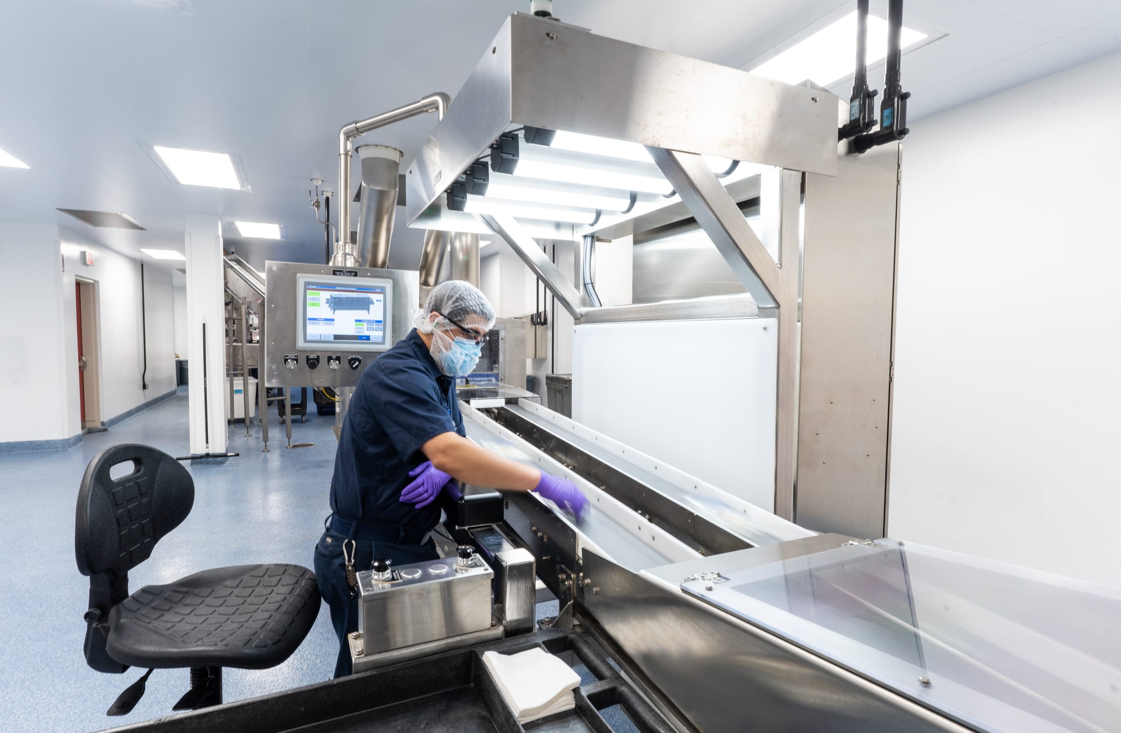 A technician wearing clean room attire including a hairnet, face mask, blue shirt, and purple gloves operates sophisticated medical device manufacturing equipment in a BSA-designed facility. The worker is operating a stainless steel processing machine with a digital control panel displaying operational parameters, while seated at a black ergonomic chair. The clean room features bright white walls, recessed lighting, blue flooring, and visible hallway access points with strategic lighting. The specialized equipment includes transparent guard panels, overhead mechanical components with illumination, and precise manufacturing controls for maintaining the sterile production environment required for medical device fabrication.