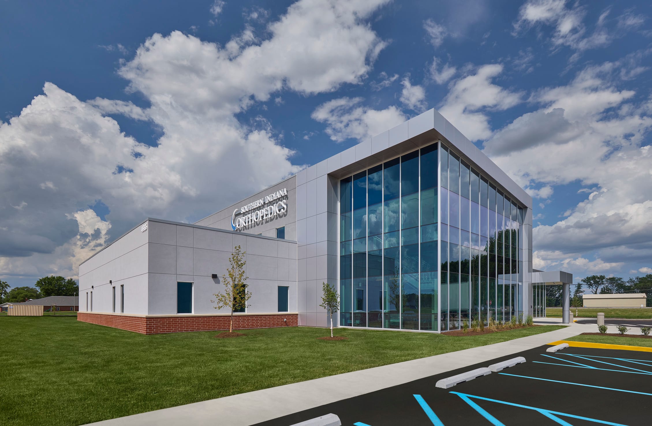 The exterior of Southern Indiana Orthopedics in Columbus, IN, presents a sleek and modern healthcare facility designed with clarity, precision, and a patient-first mindset. The architecture features a clean palette of light gray panels and red brick accents at the base, establishing a grounded, welcoming presence. A striking glass volume at the building’s entrance anchors the design, flooding the interior with natural light while offering transparency and visual connection to the surrounding landscape. Sharp lines, a refined material palette, and accessible design elements reflect a commitment to wellness, innovation, and community care.