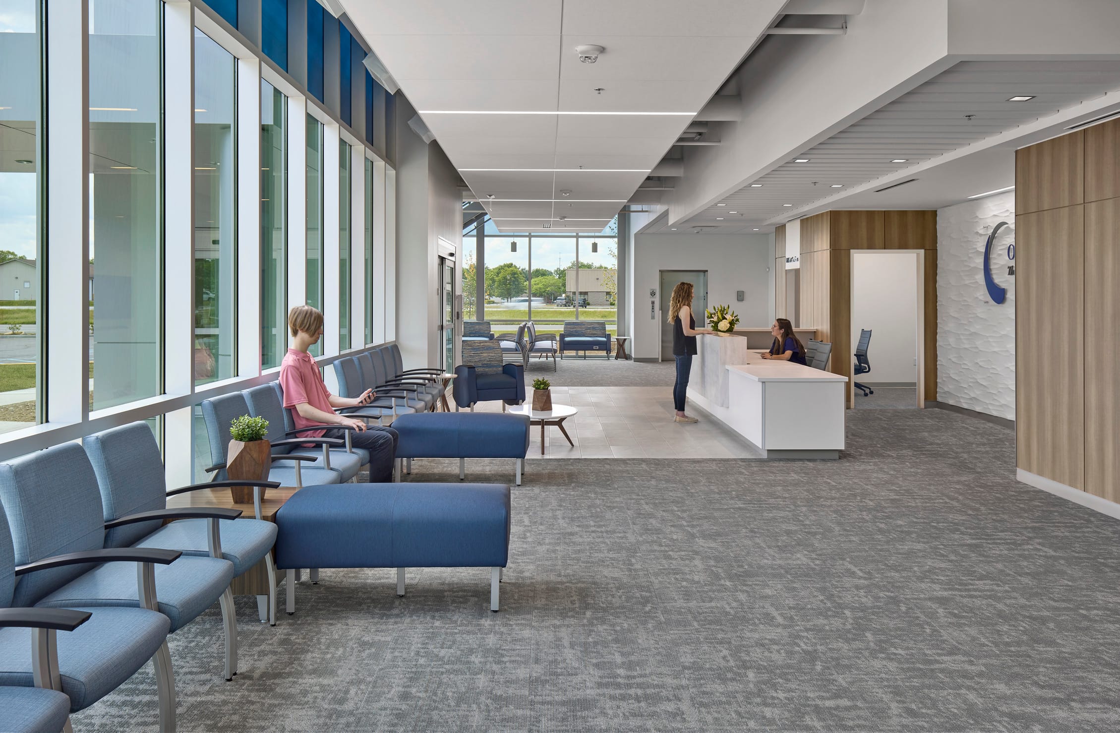 The image shows the modern reception and waiting area of Southern Indiana Orthopedics, featuring a bright, open design with floor-to-ceiling windows along the left wall that bring in natural light and provide views of the exterior. A person in a pink shirt sits in the waiting area, which is furnished with comfortable blue and gray seating including chairs and ottomans arranged in small groupings. The space uses a calming color palette of blues, grays, and natural wood tones. A clean white reception desk is positioned on the right side where a staff member is assisting a visitor. Warm wood accent walls and textured panels add depth to the space, and small potted plants provide touches of greenery. The overall design creates a welcoming, contemporary healthcare environment that feels more like a hotel lobby than a traditional medical waiting room.